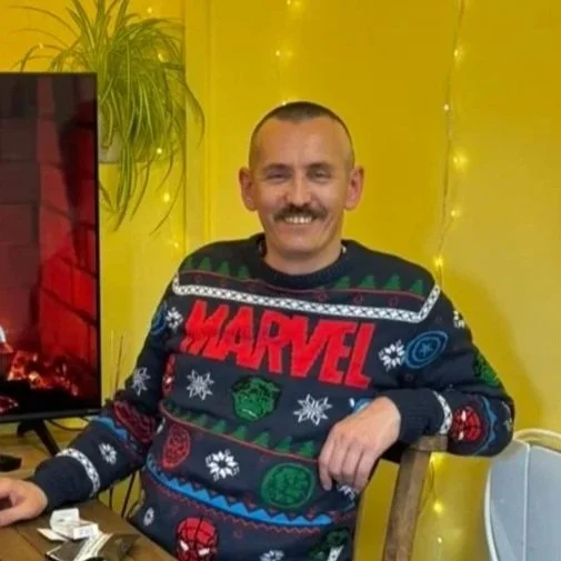 Man with a mustache smiling, wearing a Marvel-themed Christmas sweater, sitting at a table in a decorated room with yellow walls and string lights.