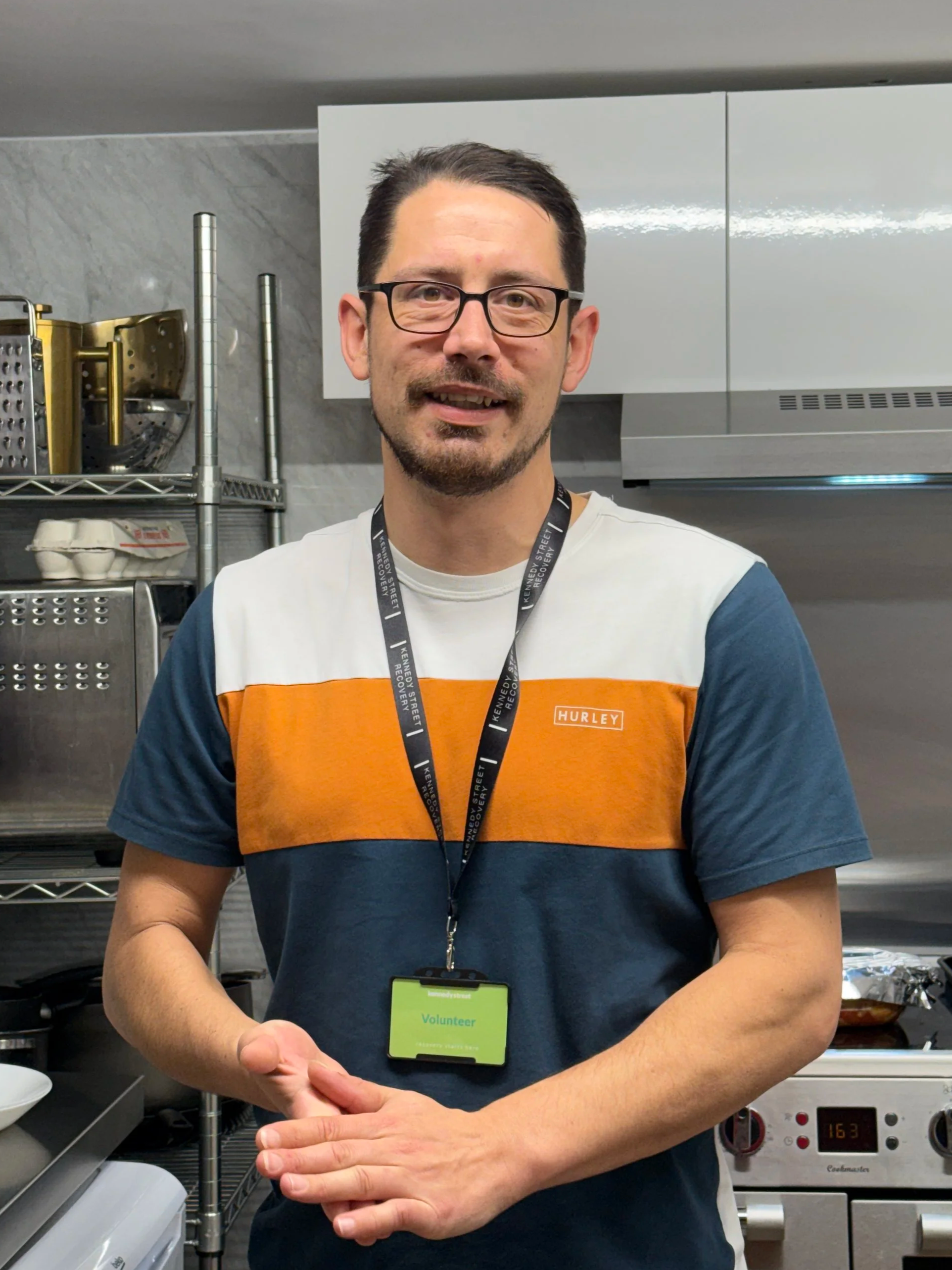 A man wearing glasses and a Harleys t-shirt, standing in a kitchen with industrial kitchen equipment in the background. He is smiling, with a volunteer badge hanging around his neck. The man appears to be doing a hand gesture.