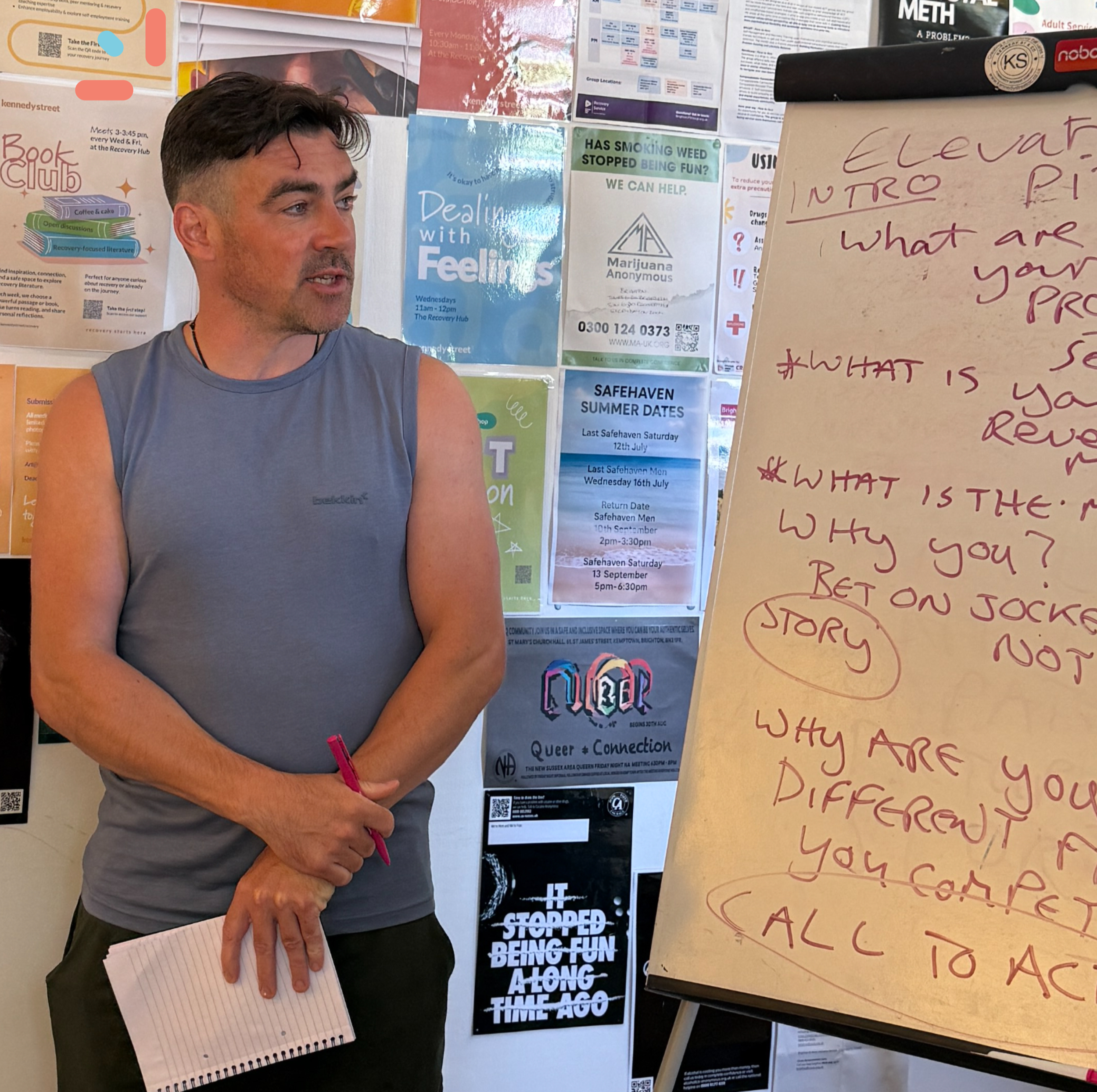 A man holding a pink pen and a notebook stands next to a handwritten poster board with red ink. The background features various colorful posters on a wall, including ones related to recovery, mental health, and drug awareness.