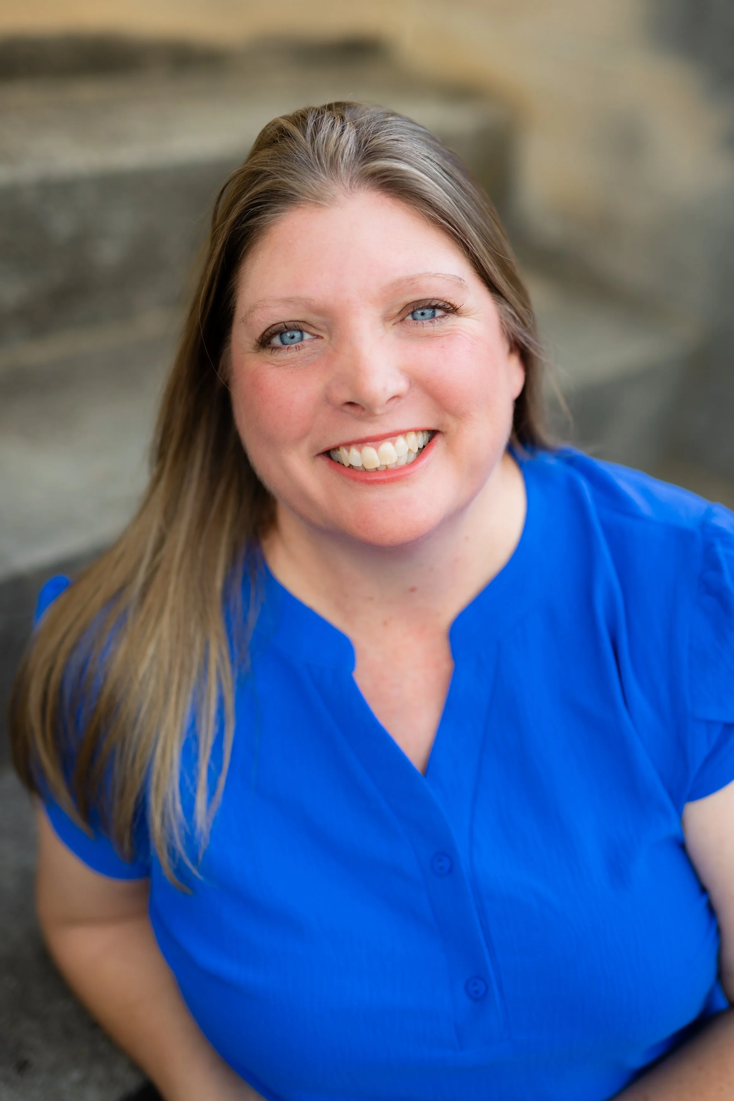 Close-up of a smiling woman with long, light brown hair and blue eyes, wearing a blue shirt, sitting outdoors.