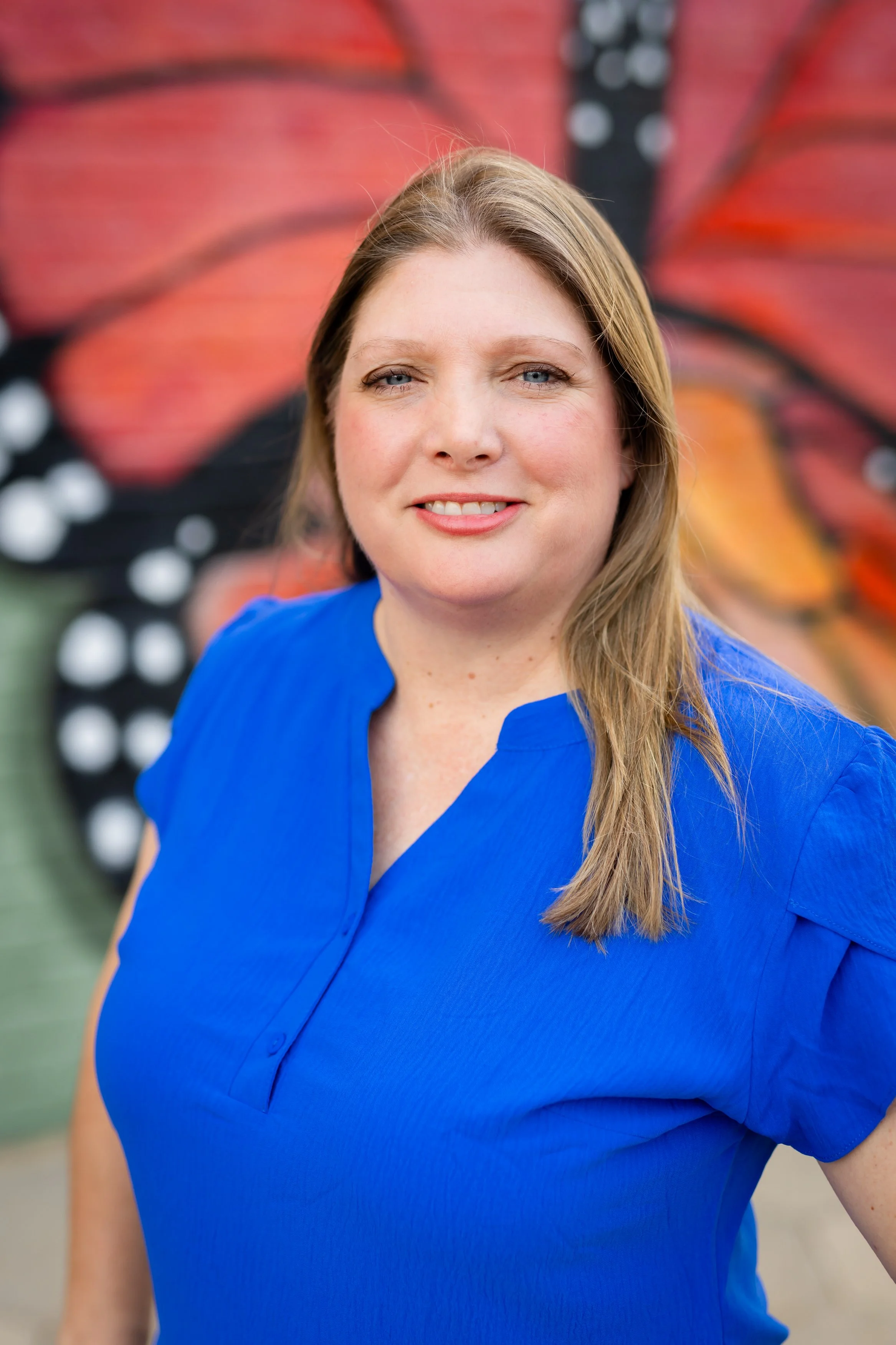 Close-up of a woman with long light brown hair, blue eyes, and light skin wearing a bright blue blouse, standing in front of a large butterfly mural with orange, black, and white wing patterns.