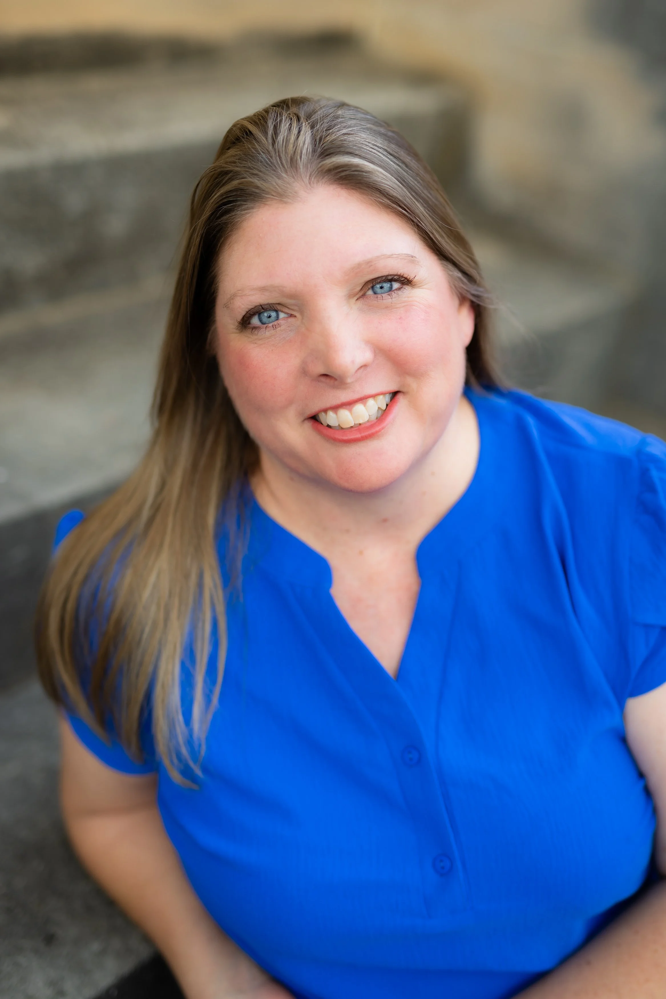 Close-up of a woman with long blonde hair, blue eyes, and a bright smile, wearing a blue shirt, outdoors.
