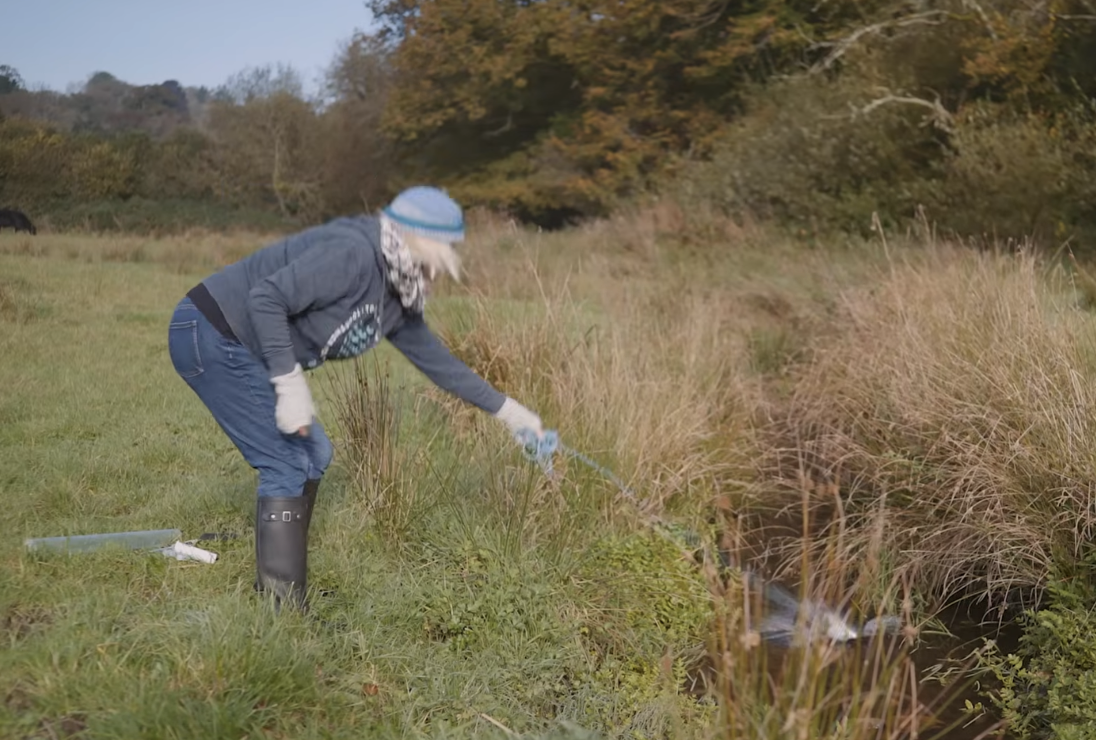 Vicky Whitworth doing water testing, bending over near a small stream in a field with tall grass and trees with autumn foliage in the background.