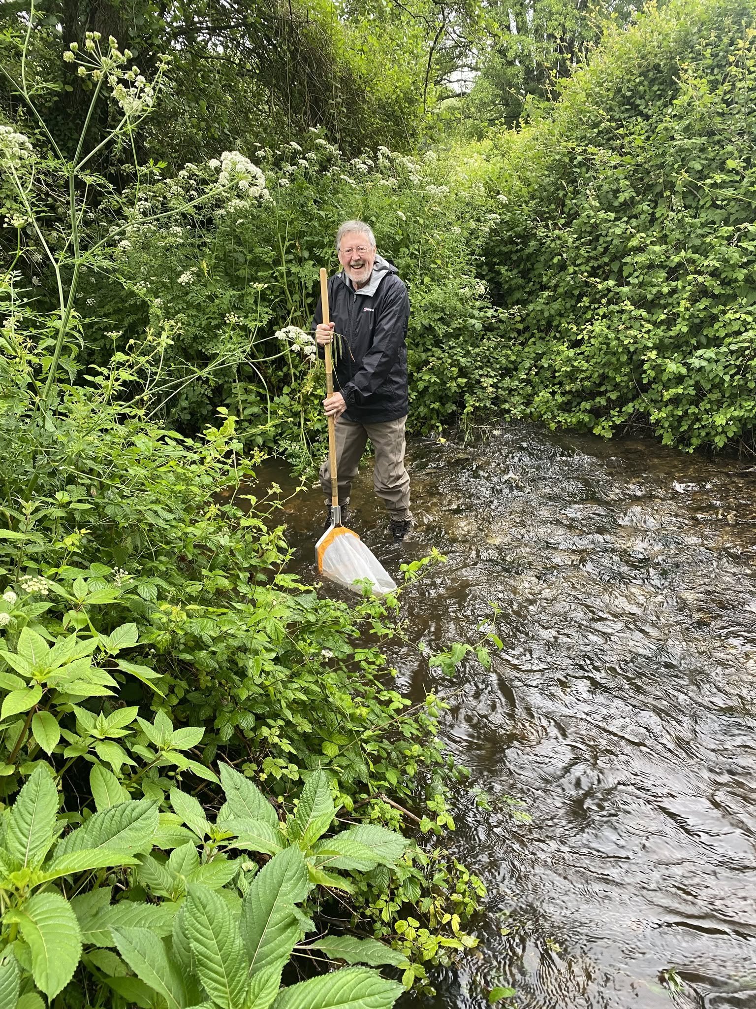 A man standing in a creek surrounded by dense green foliage, wearing a black rain jacket and beige waders, holding a net, and smiling.
