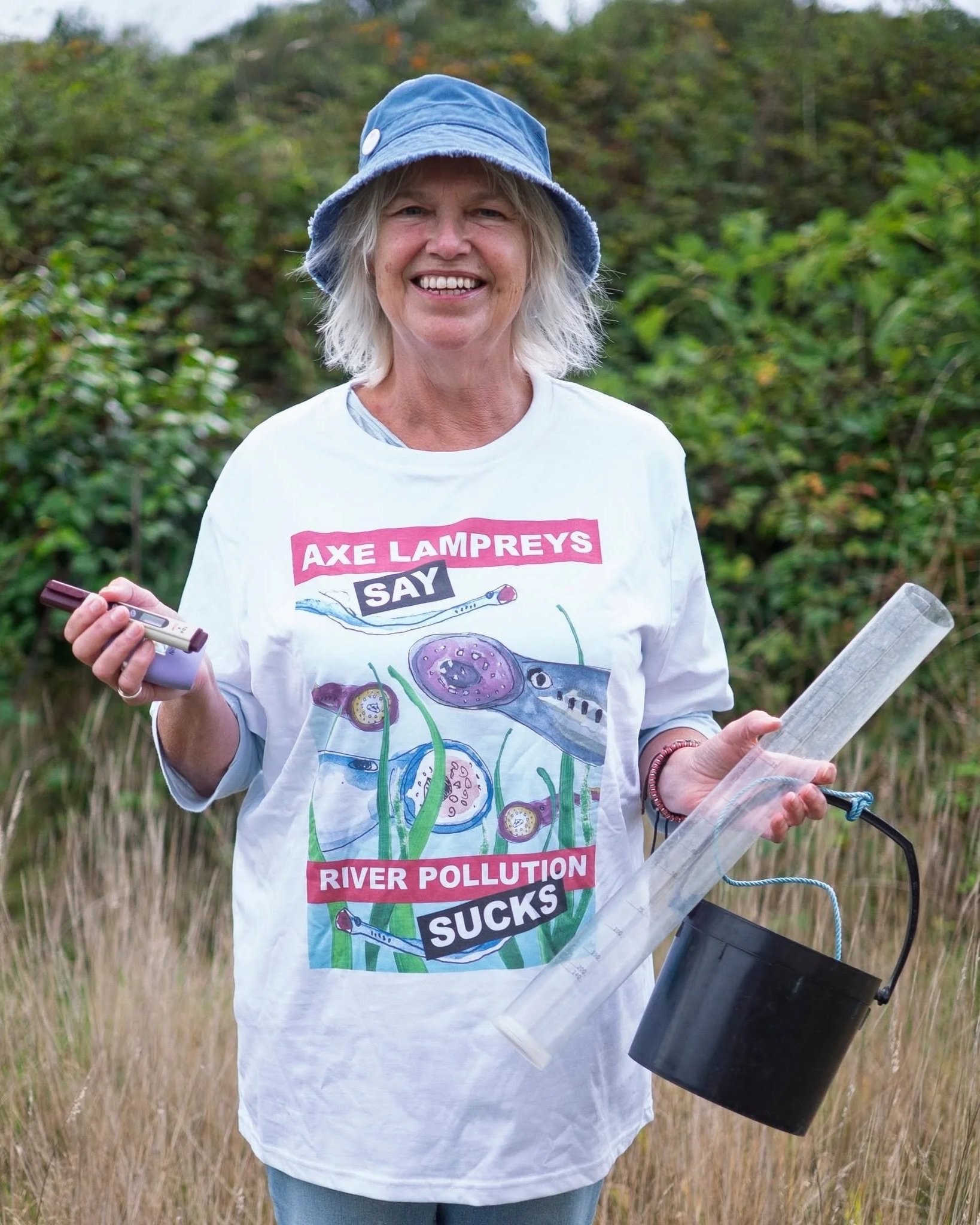 Vicky Whitworth, holding her water testing equipment and Axe Lampreys say no to water pollution message t-shirt, standing outdoors in a natural setting with green bushes and tall grass.