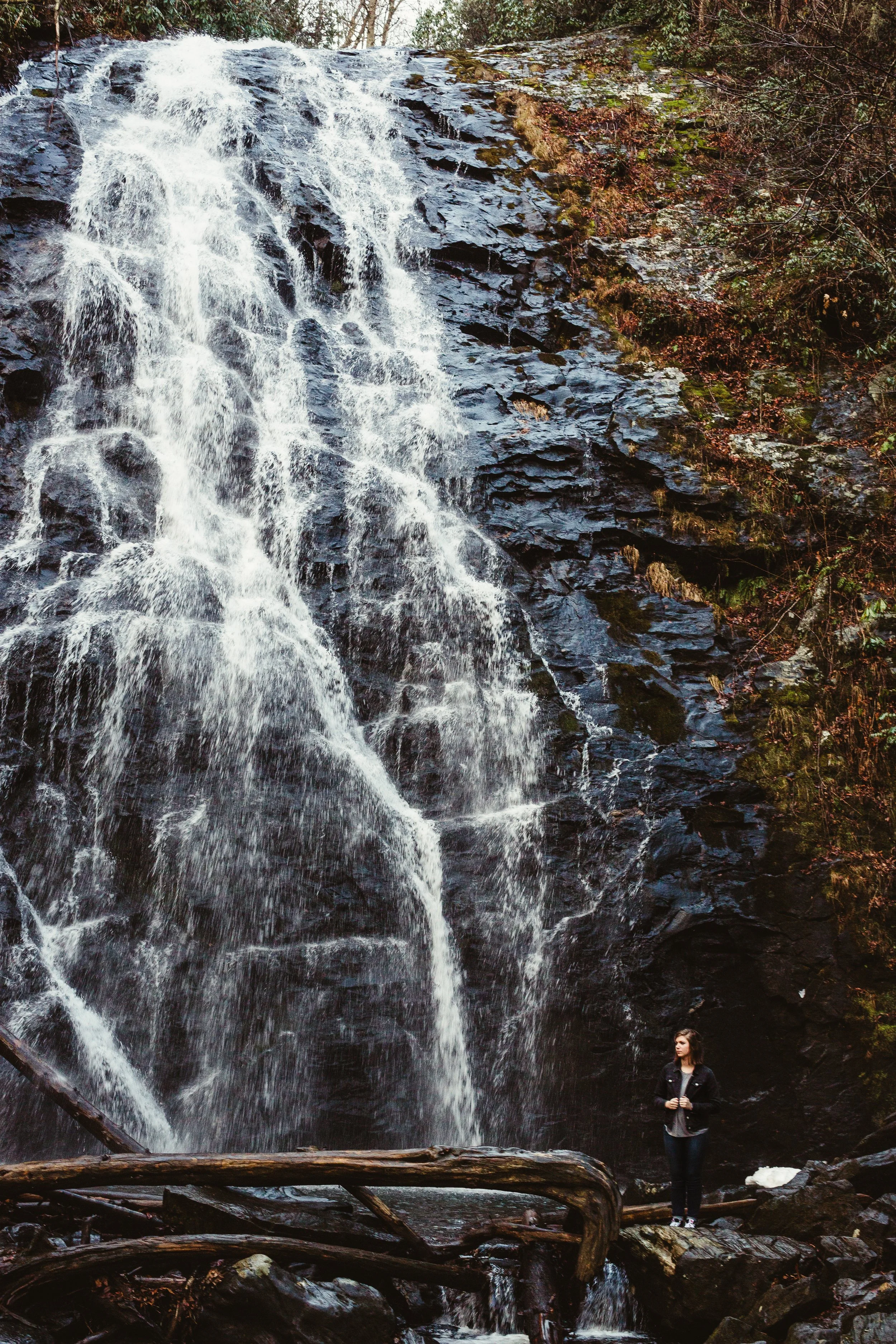 A woman standing on rocks near a tall waterfall in a forested area.