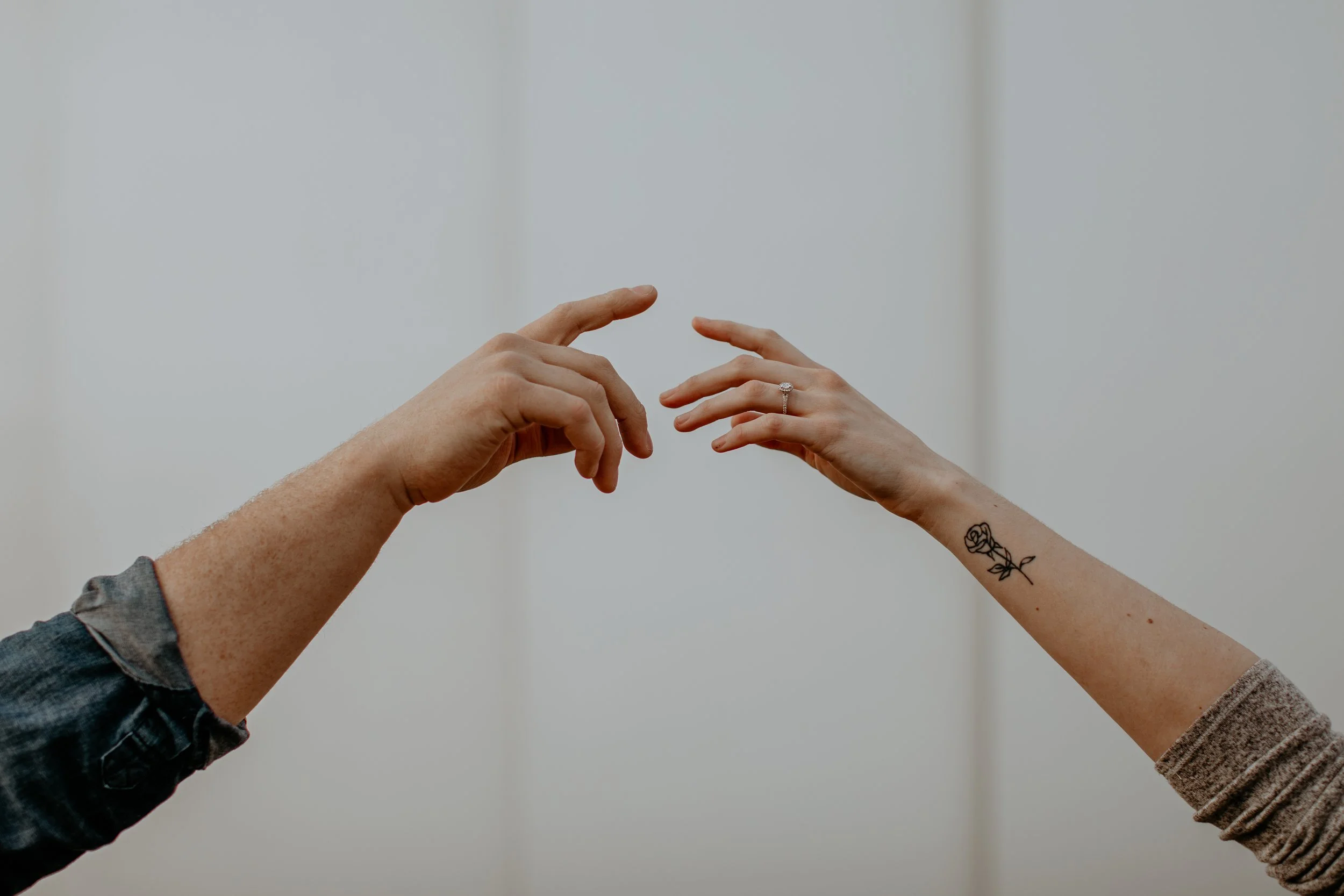 Two hands reaching towards each other with fingers nearly touching, one hand has a ring and a tattoo of a rose on the forearm, the background is a plain wall.