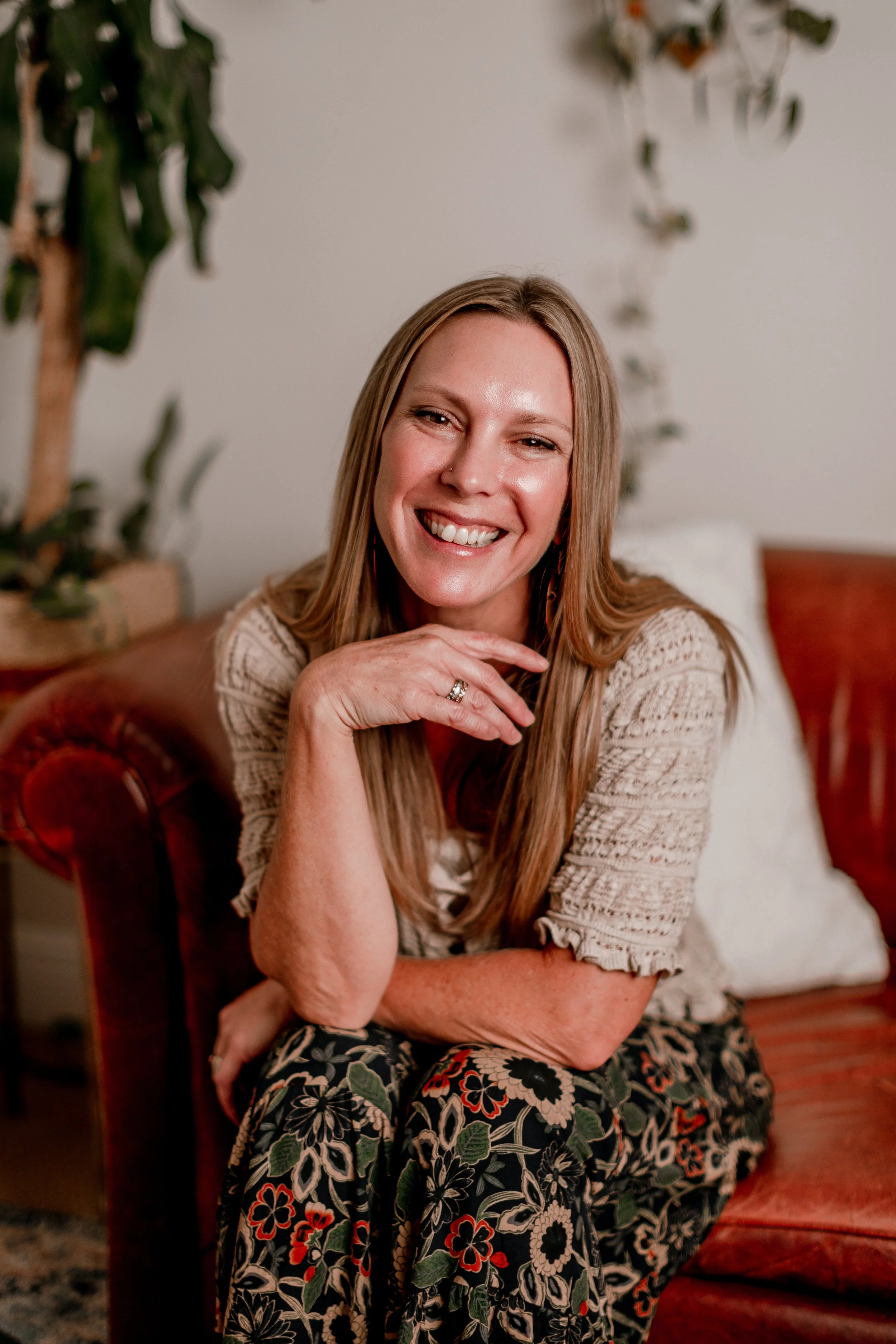 A smiling woman with long light brown hair sitting on a vintage red velvet armchair, wearing a cream-colored sweater and a floral skirt with black, red, white, and green colors, in a cozy indoor setting with plants in the background.