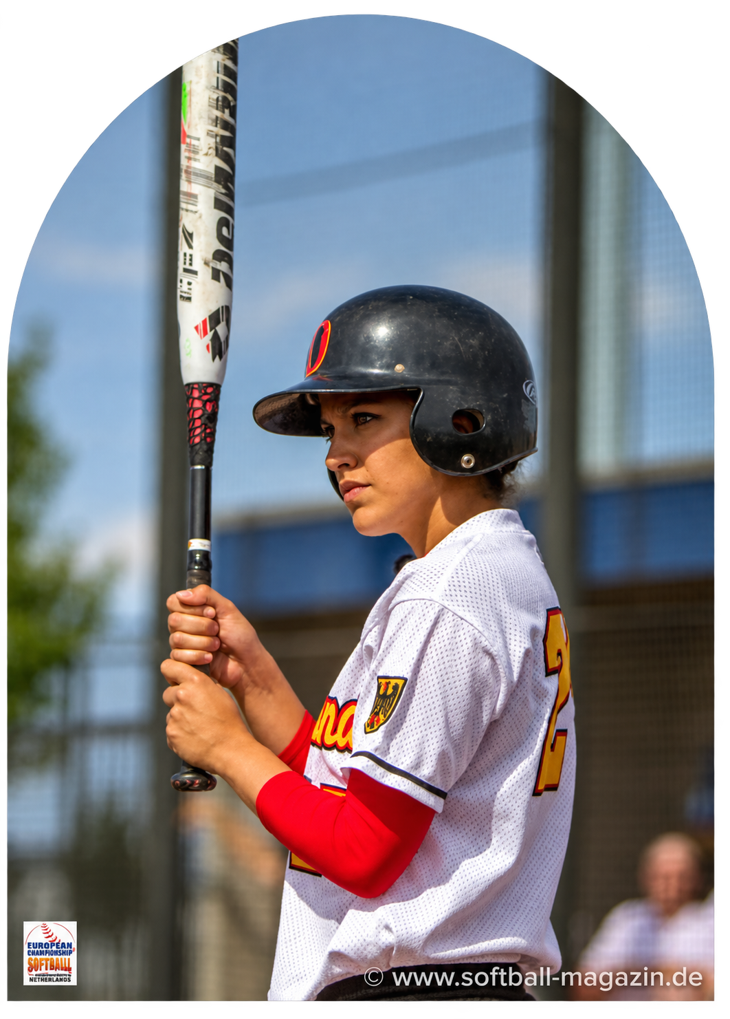 Junge Frau in Softballuniform mit Helm und Softball-Schläger auf dem Spielfeld, bereit zum Schlag, europäische Softballmeisterschaft, Niederlande, moderne Sportkleidung, klare Himmel im Hintergrund.