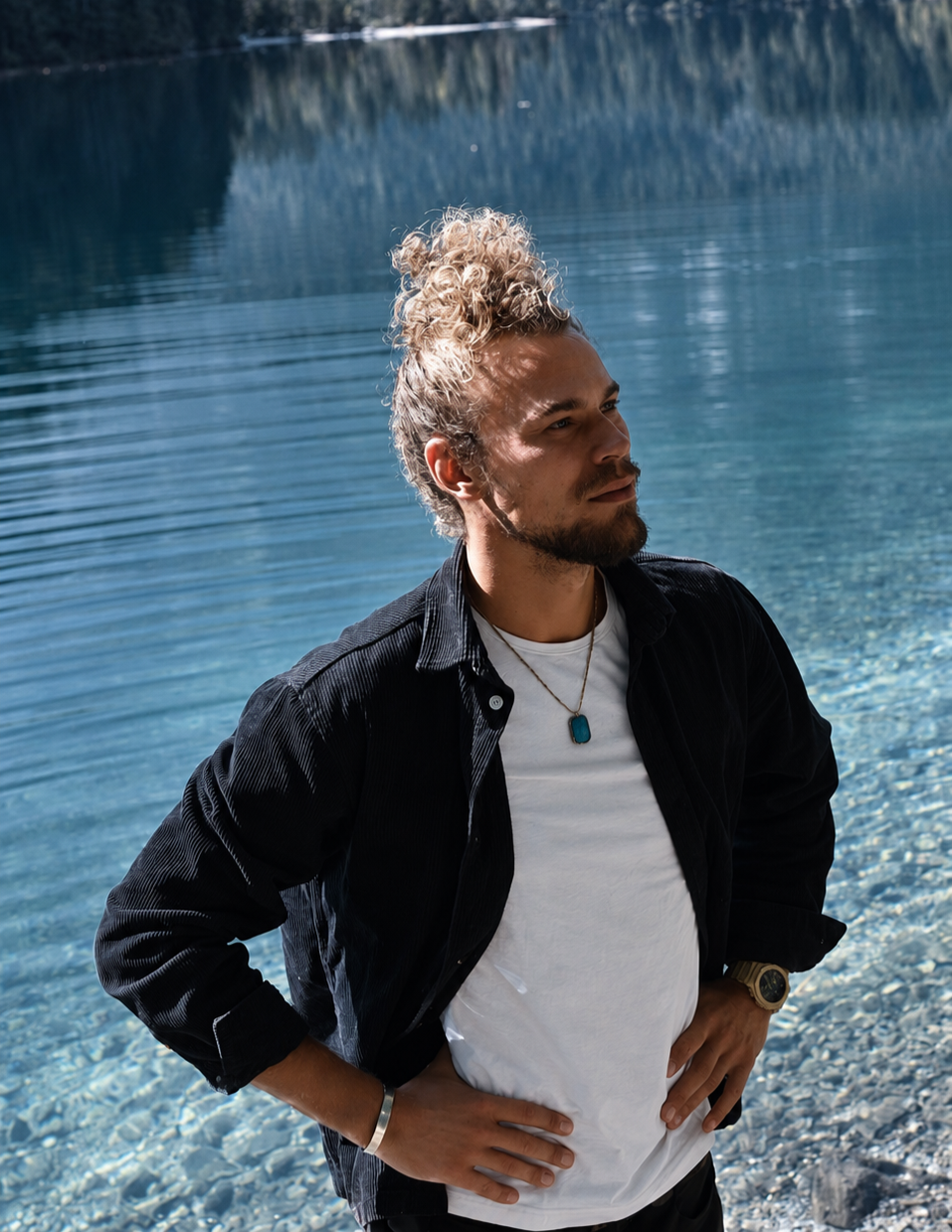 Junger Mann mit blond lockigem Haar, dunkler Jacke, weißem T-Shirt, am Ufer eines klaren Sees, mit Wasser und Bäumen im Hintergrund.