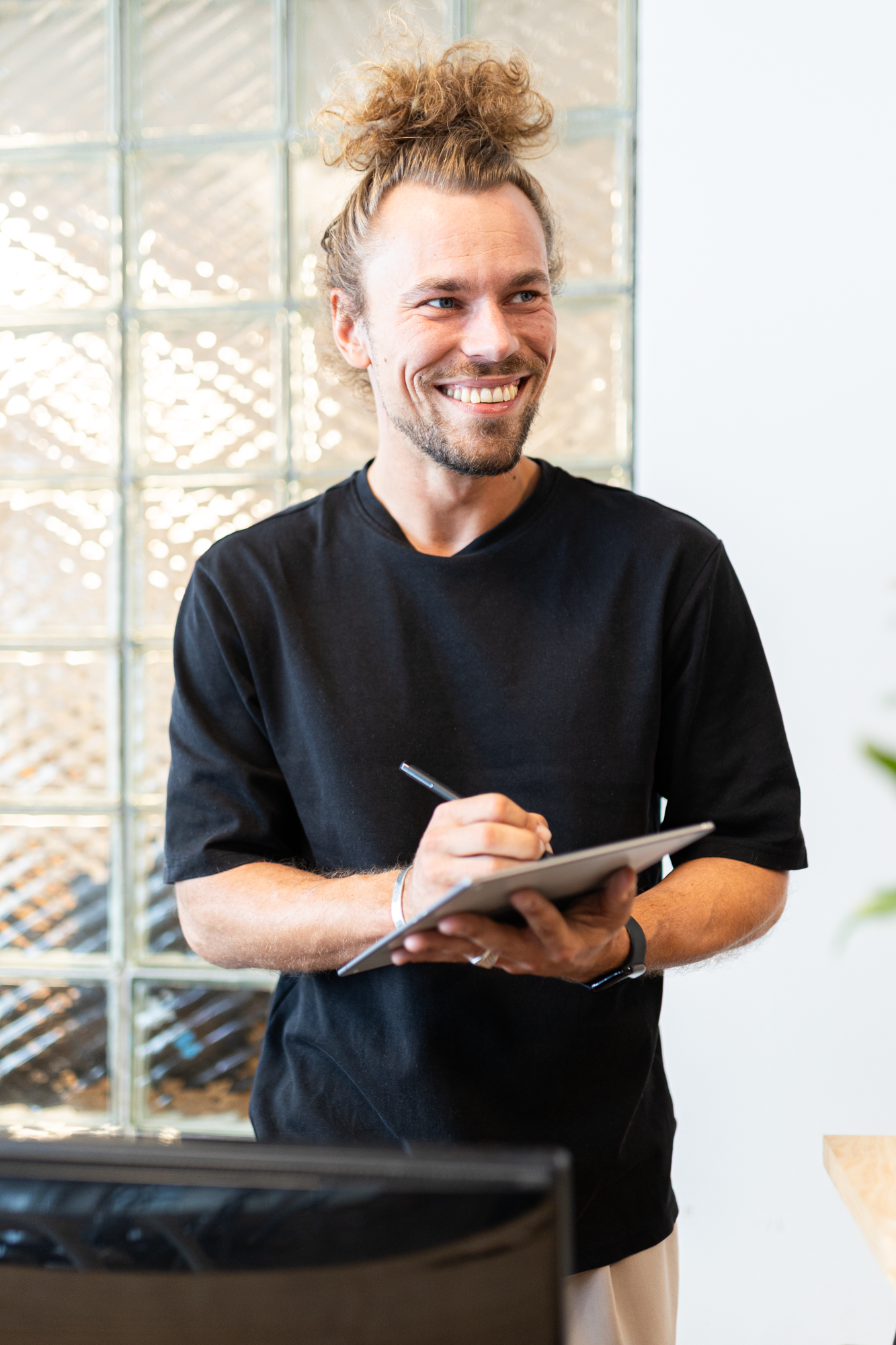 Ein Mann mit Locken, Bart und einem lockeren Haar Dutt trägt ein schwarzes T-Shirt und hält einen Notizblock mit Stift in den Händen, während er lächelnd in einem Raum steht, vor einem Fenster mit Glassteinen.