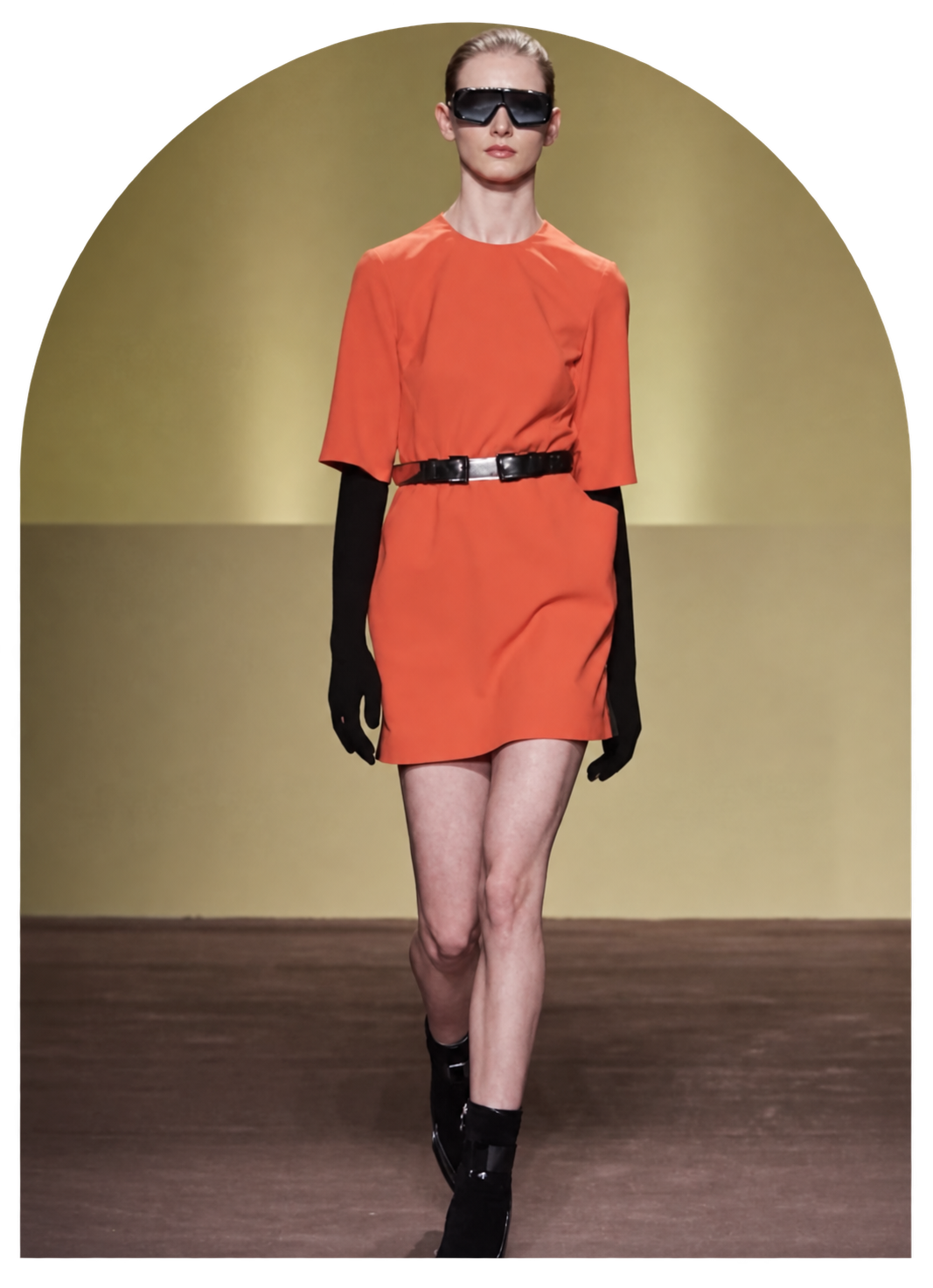 Eine Modenschau mit einer weiblichen Laufsteg-Model, die ein rotes Kleid trägt, schwarze Handschuhe, Sonnenbrille und schwarze Schuhe. Der Hintergrund ist gelb.