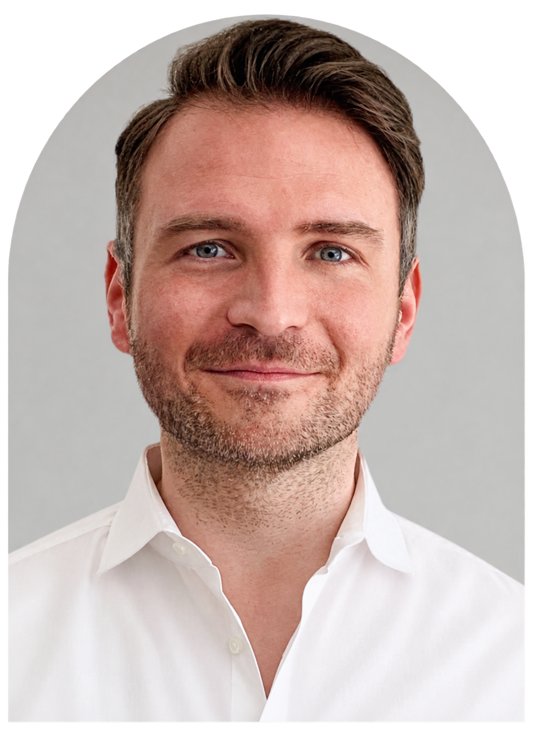 Ein Mann mit braunen Haaren, blauen Augen, Bart und weißem Hemd vor grau Hintergrund.