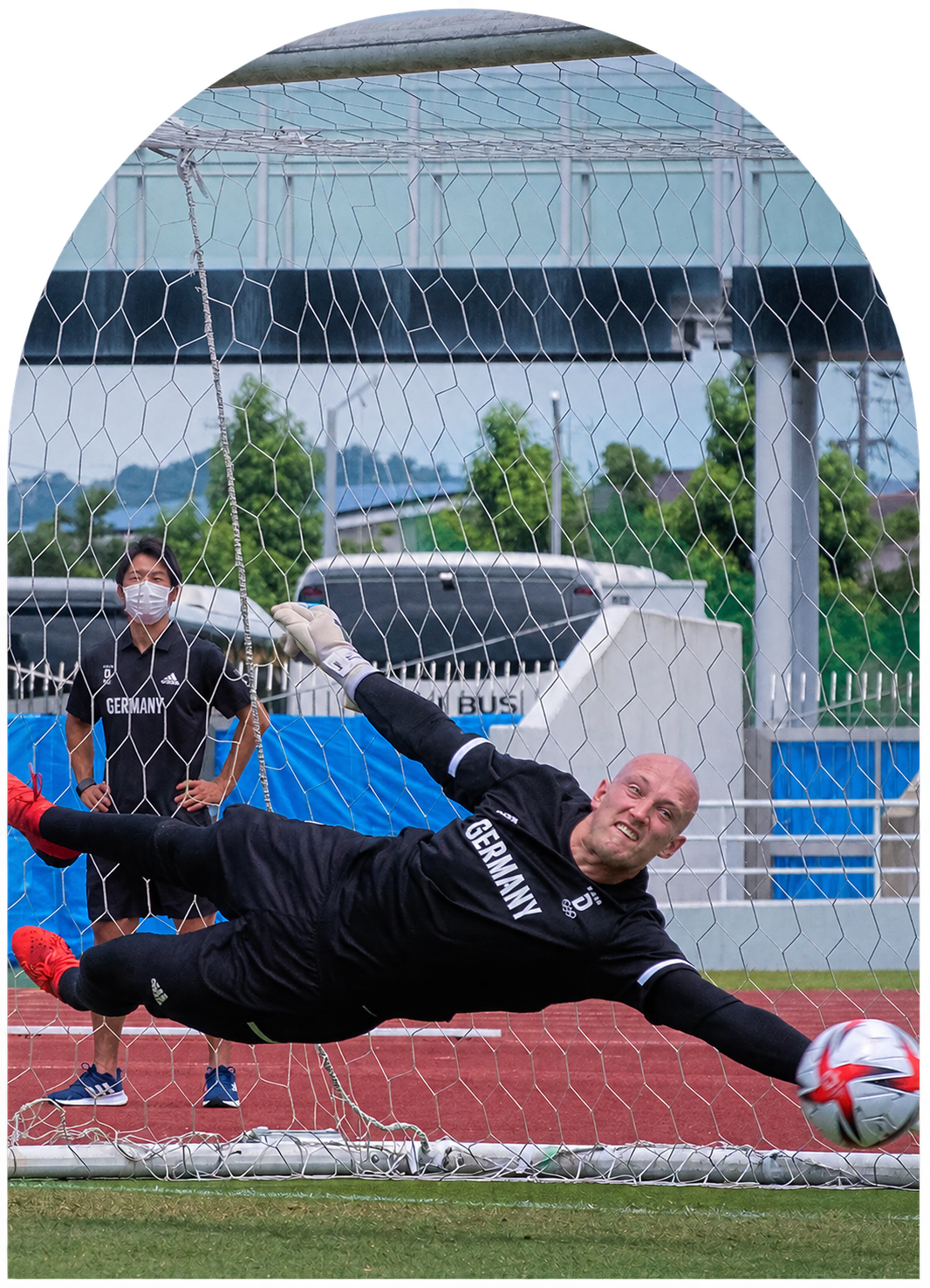 Ein Fußballspieler im Tor, der die Kugel auf dem Spielfeld hält, während eine Person im Hintergrund zuschaut. Das Bild zeigt eine Spielsituation bei einem Football-Training oder -Spiel.