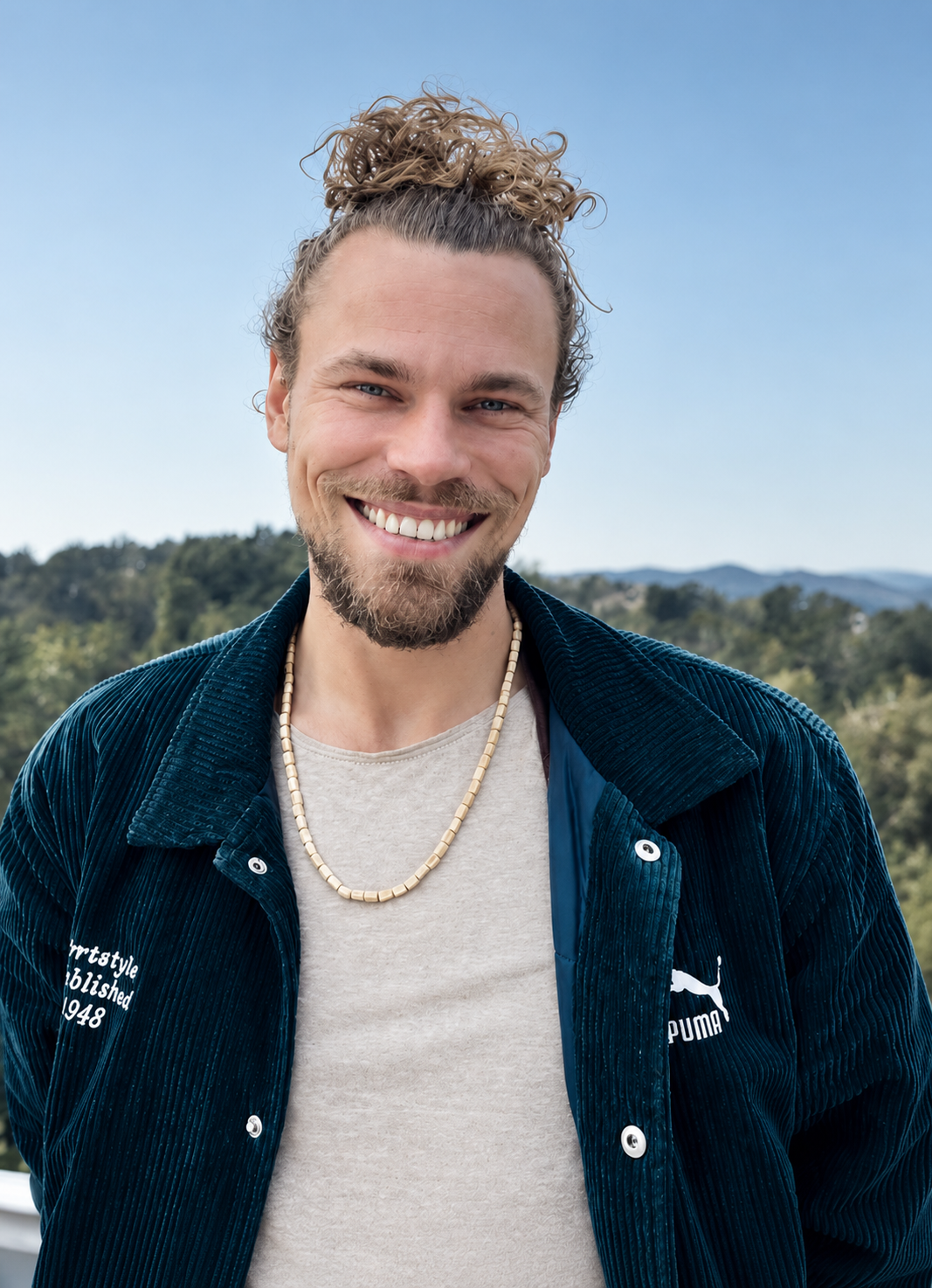 Ein lächelnder Mann mit langem, lockigem Haar, Bart und Schnurrbart, trägt eine beige Halskette, eine graue T-Shirt und eine dunkelblaue, strukturierte Jacke mit Puma-Logo, vor einer grünen Landschaft unter blauem Himmel.