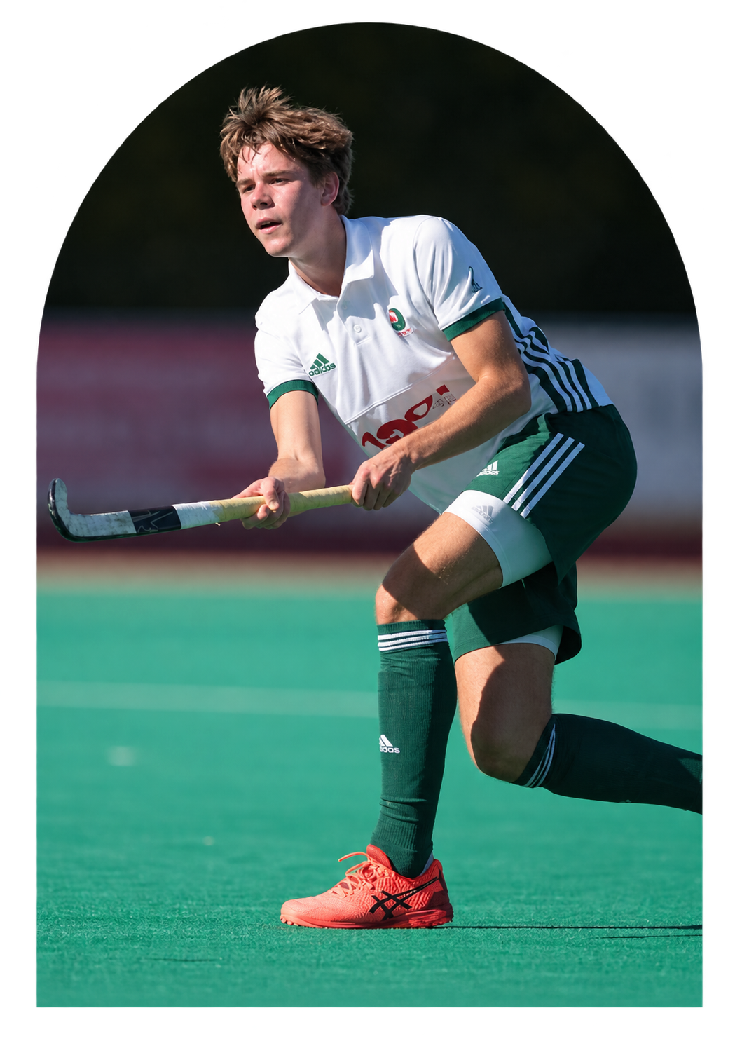 Mann spielt Feldhockey auf einem grünen Platz, trägt ein weißes Trikot, grüne Shorts und rote Sportschuhe.