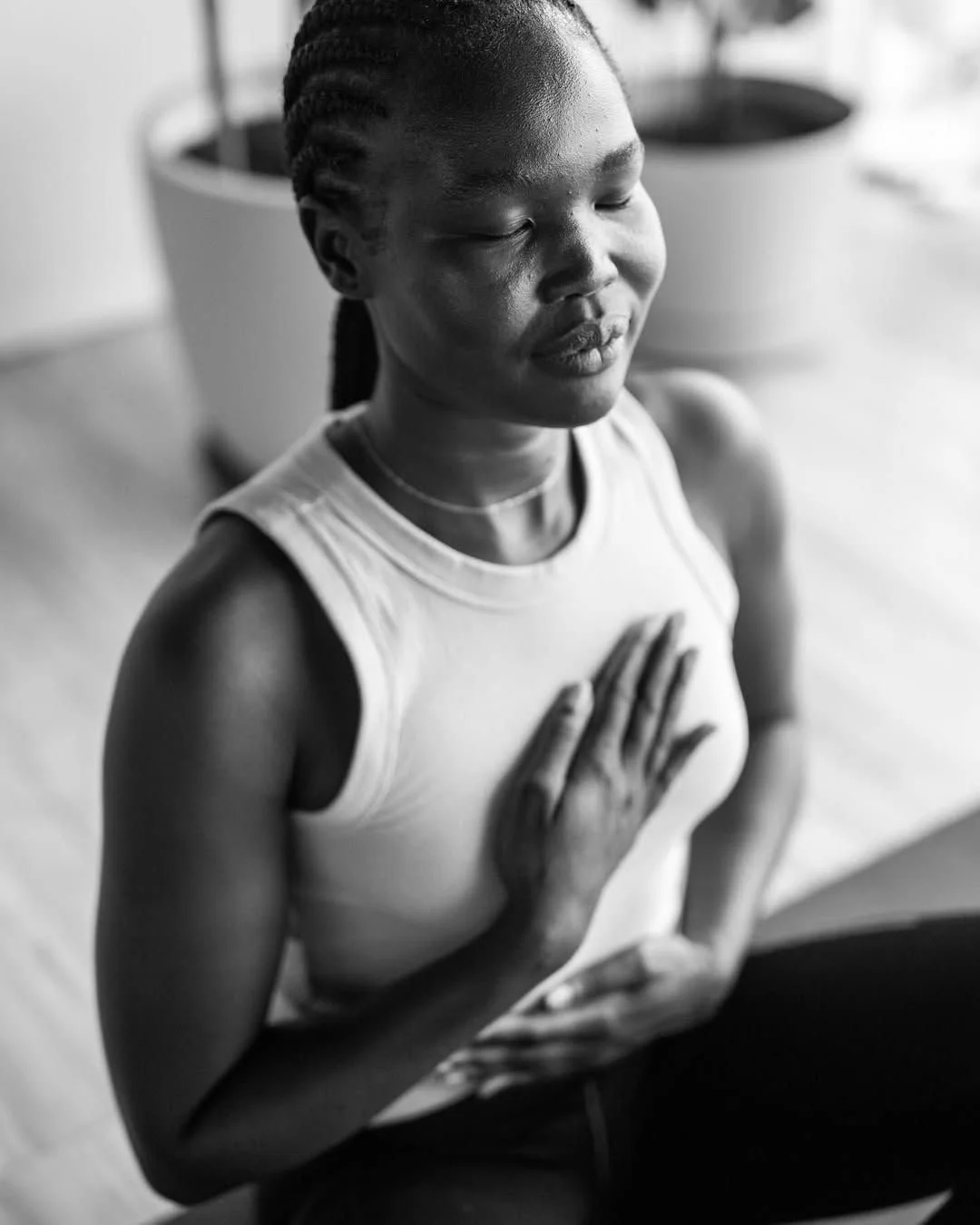 Eine junge Frau macht Yoga und hält die Hand aufs Herz, im Innenraum, die Augen geschlossen, entspannter Gesichtsausdruck, schwarz-weiß Fotografie.