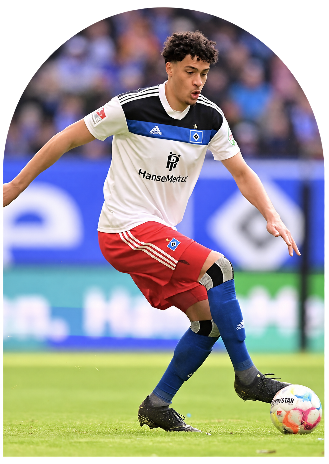 Fußballspieler in weißen Trikot mit schwarzen, blauen und roten Details, rot-weiße Shorts, blaue Socken und schwarze Schuhe, während er einen Ball auf einem Feld spielt.