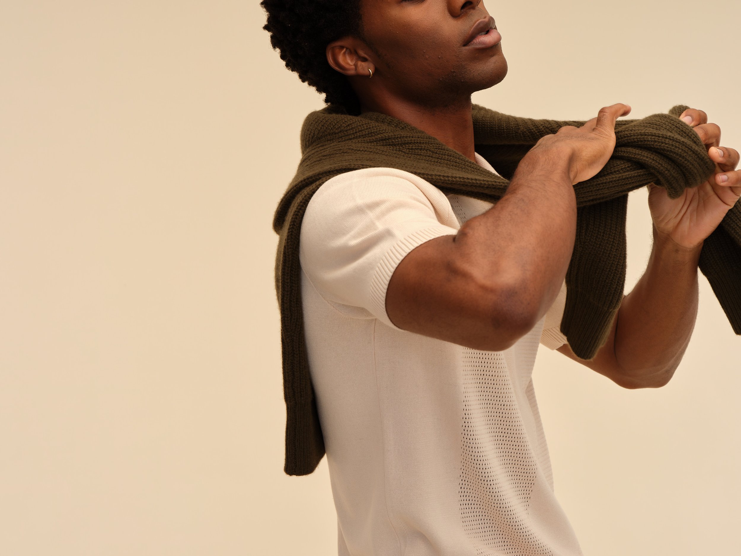 A person with dark skin and short curly hair is holding a brown sweater over his shoulders, wearing a white short-sleeve shirt, and standing against a beige background.