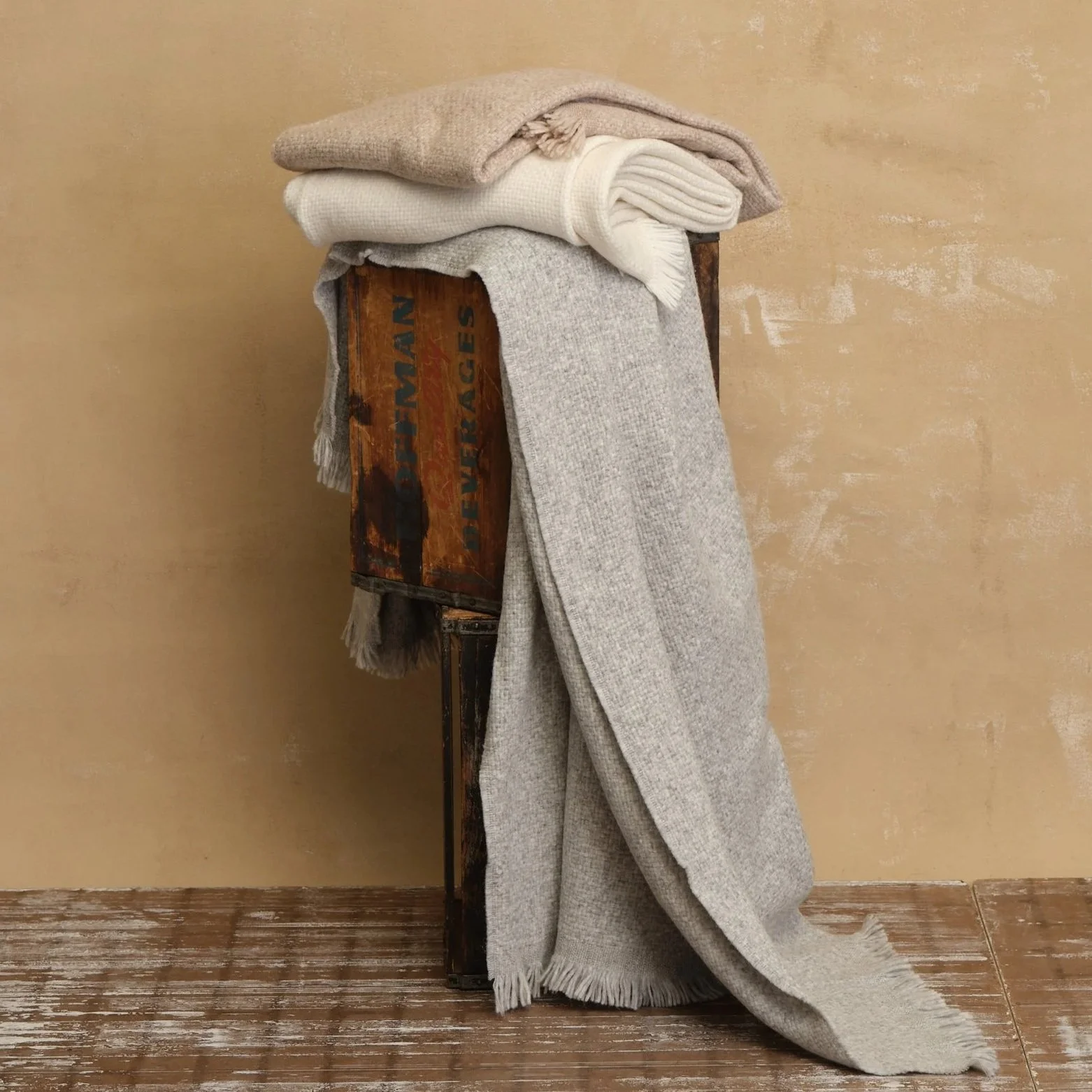 A stack of folded scarves and blankets resting on a vintage wooden crate against a beige wall.