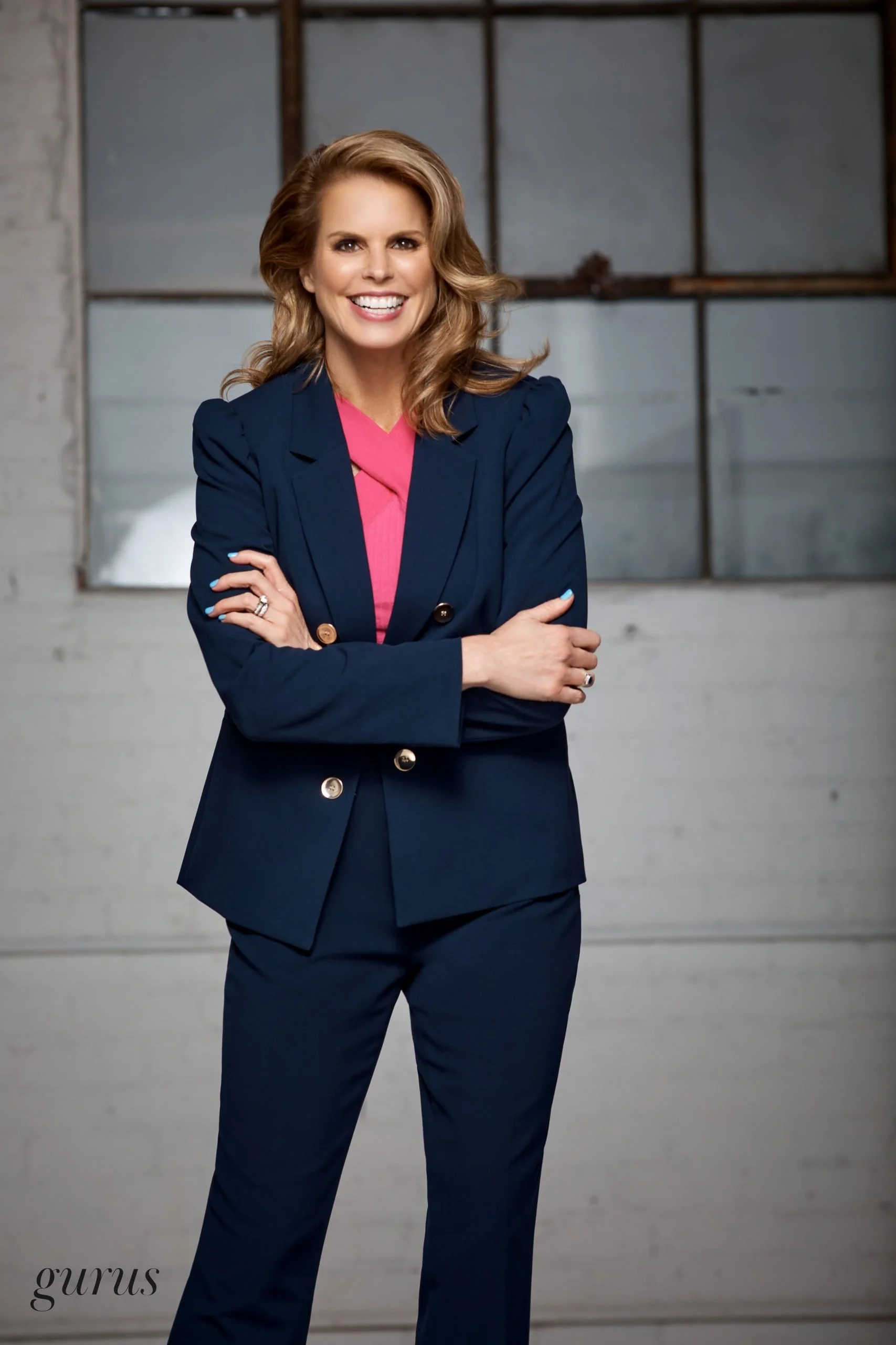 A woman with wavy blonde hair smiling, standing in front of a large industrial-style window, wearing a navy blazer and matching pants, with a pink top underneath.