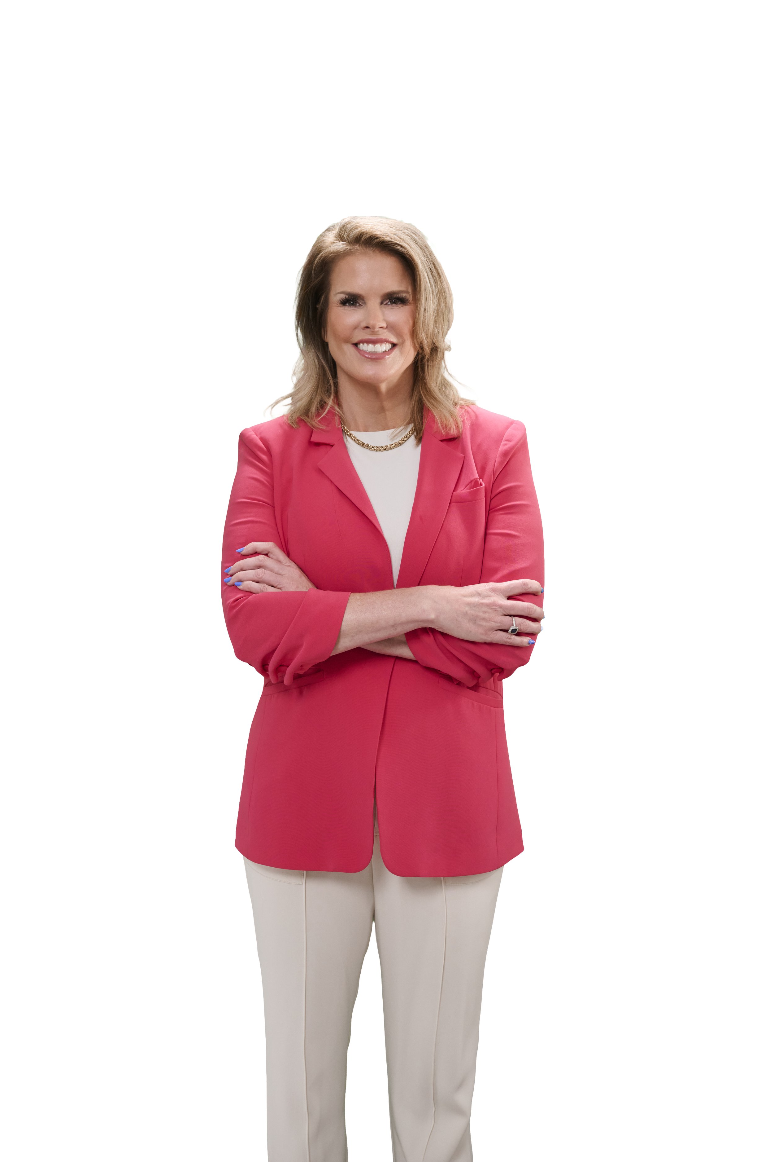 A woman with shoulder-length blonde hair, smiling, wearing a pink blazer over a white shirt, crossing her arms, paired with beige pants, standing against a plain white background.