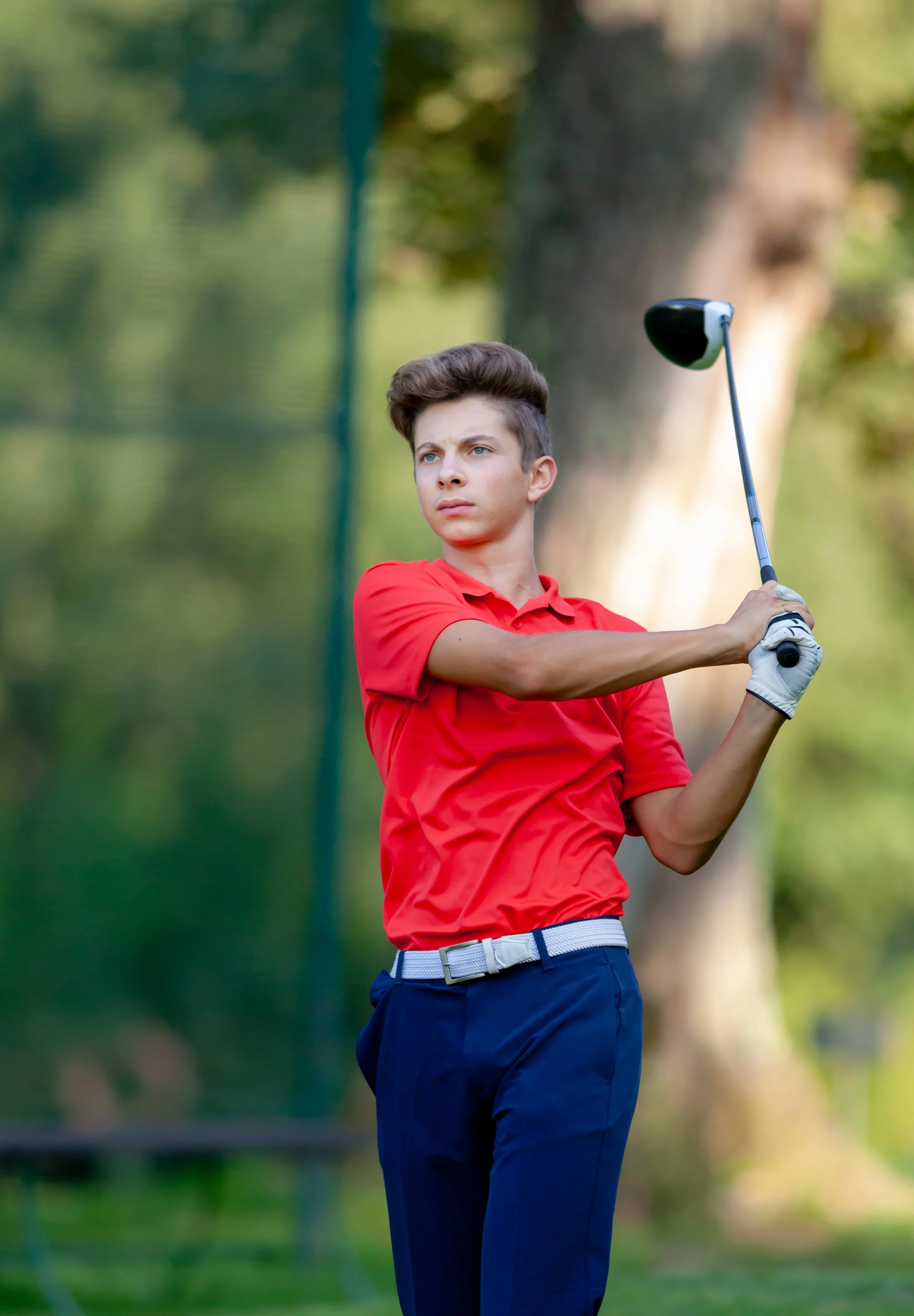 A young male golfer wearing a red polo shirt, navy pants, and a white belt, swinging a golf club on a golf course with trees in the background.