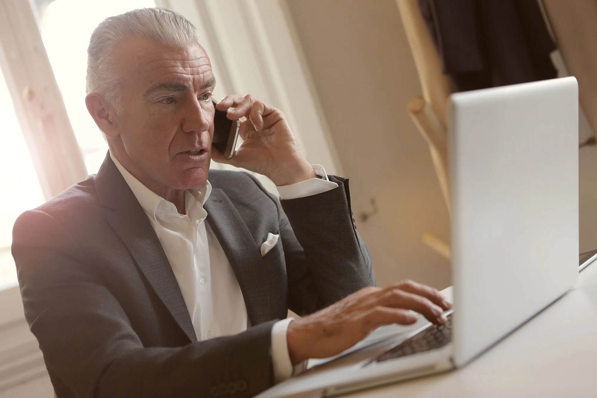 An older man with silver hair, wearing a gray suit and white shirt, is talking on a mobile phone while typing on a laptop.