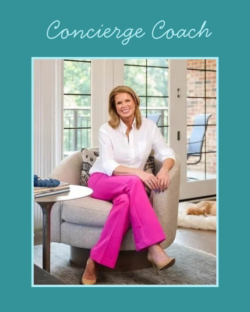 A woman sitting on a beige armchair in a bright room, smiling, wearing a white blouse and hot pink pants, with a small table beside her, a dog resting on a rug in the background, and large windows letting in natural light.