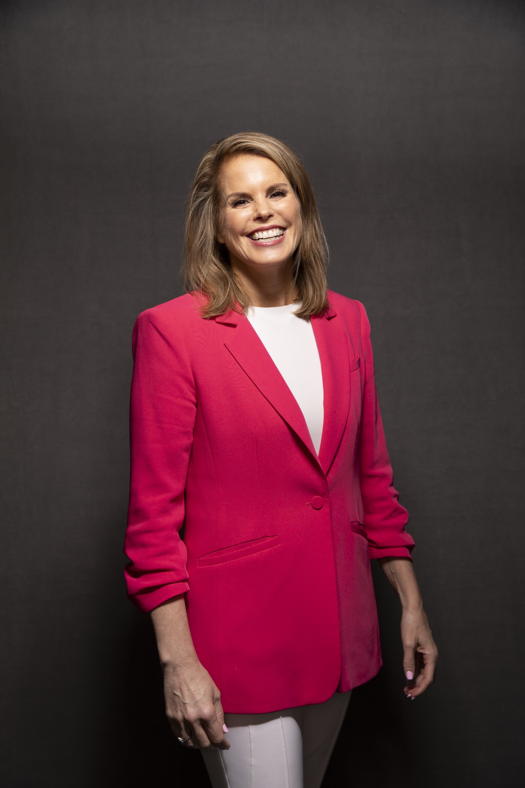 A woman smiling and wearing a bright pink blazer and white pants, standing against a dark gray background.