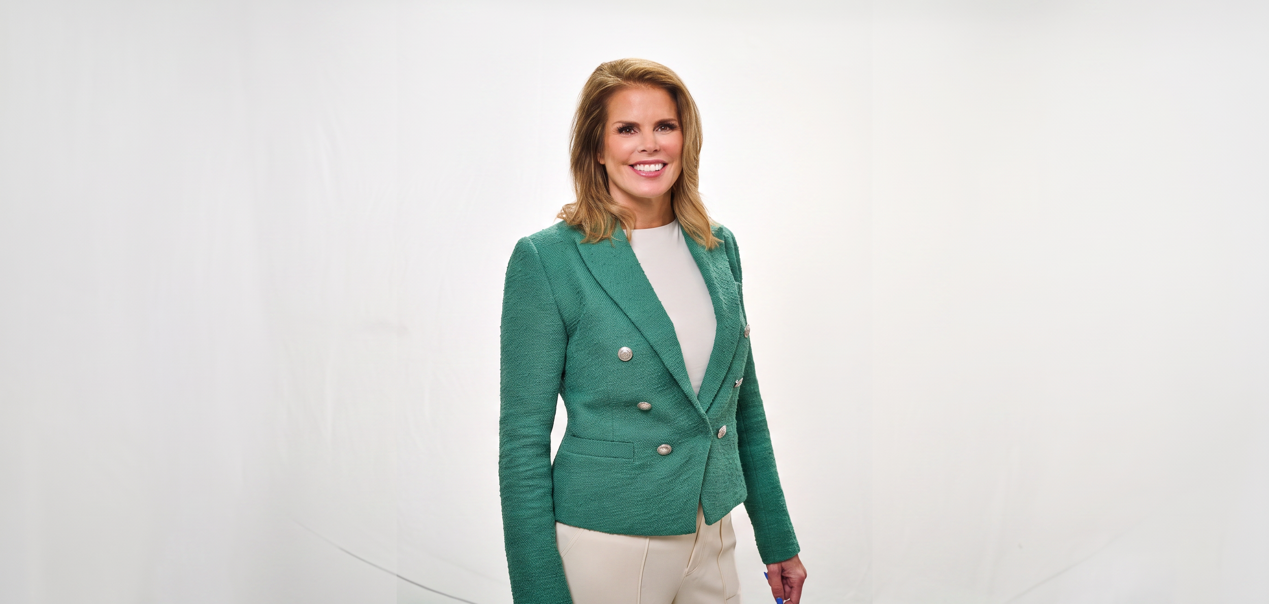 Woman wearing a green blazer and white top, standing against a white background and smiling.