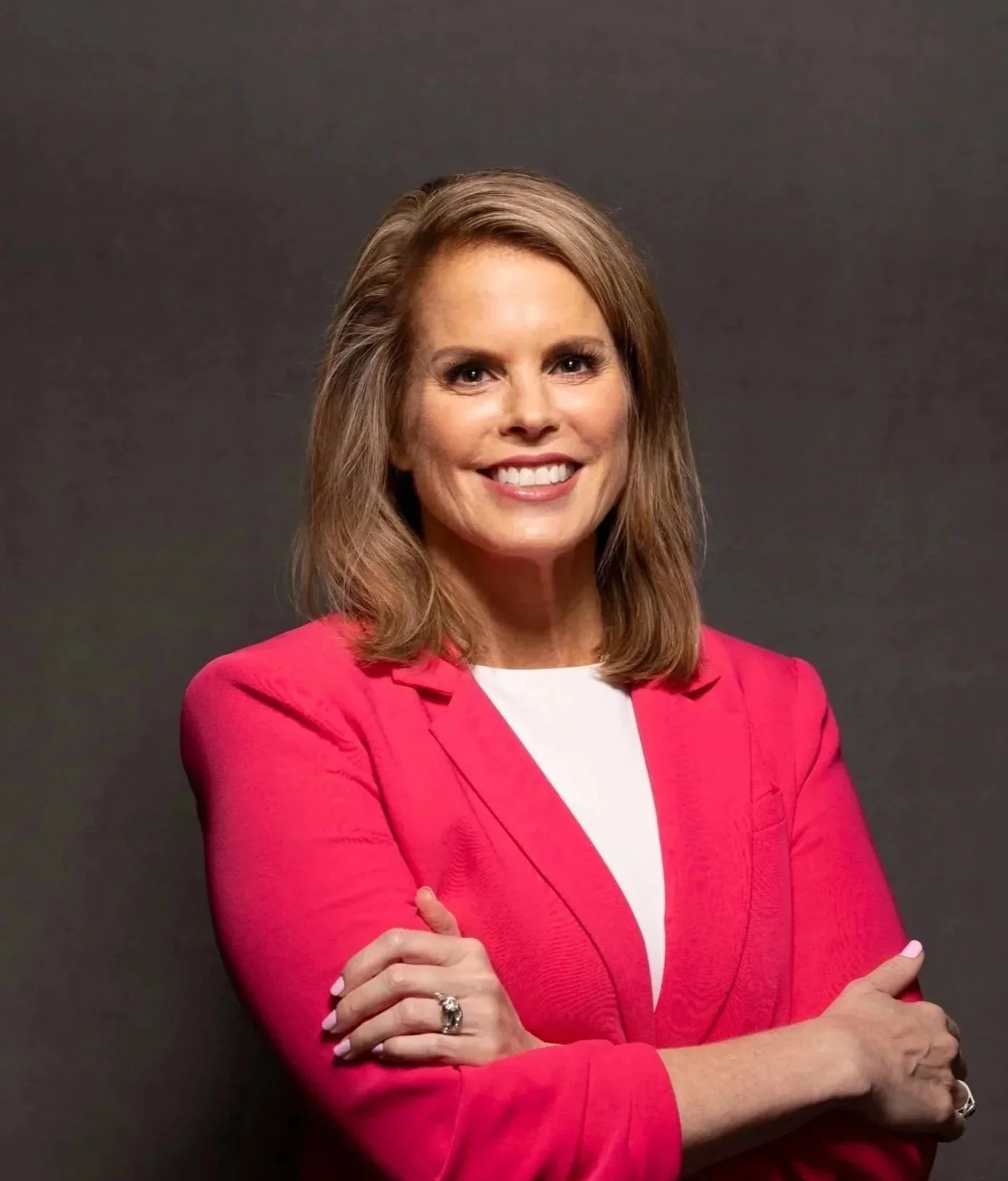 A woman with shoulder-length brown hair wearing a pink blazer and white shirt, smiling with arms crossed, against a dark background.