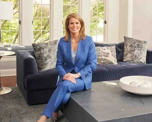 Woman sitting cross-legged on a dark blue sofa in a bright living room with large windows, dressed in a blue suit, smiling at the camera.