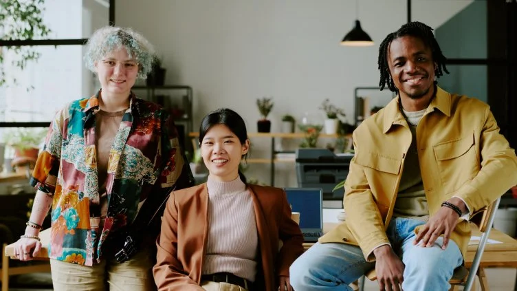 Three young adults smiling in a modern workspace. One person with curly gray hair and a colorful shirt stands on the left. A woman with straight black hair sits in the middle wearing a brown blazer. A man with dreadlocks and a yellow jacket sits on the right.