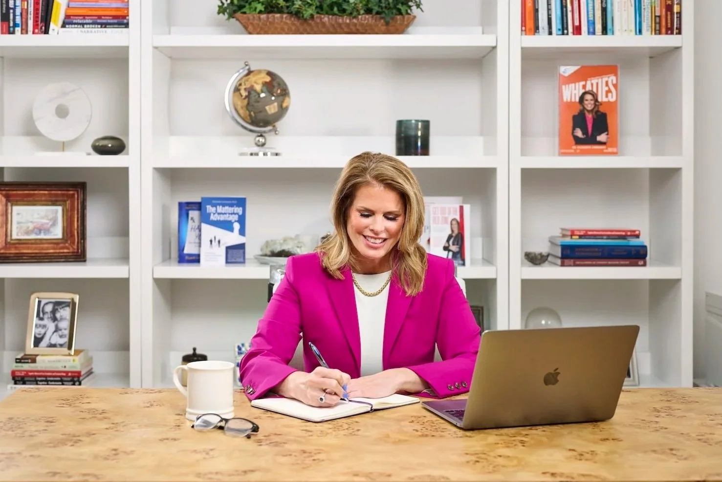 A woman in a pink blazer sitting at a desk, writing in a notebook with a silver laptop and glasses nearby, in front of white bookshelves filled with books and decorative items.
