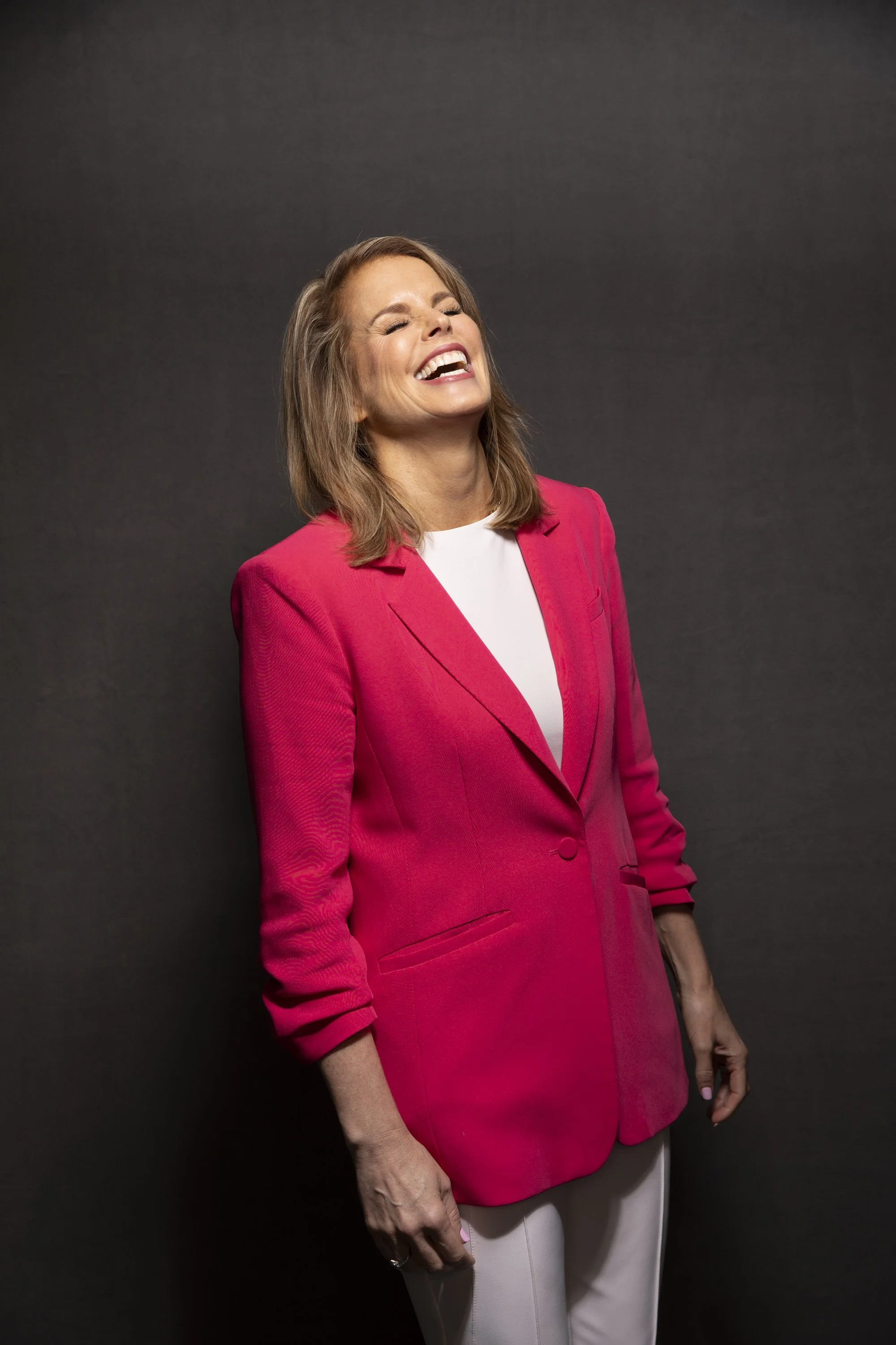 A woman with shoulder-length brown hair wearing a bright pink blazer and white pants, smiling broadly with eyes closed, standing against a plain dark background.