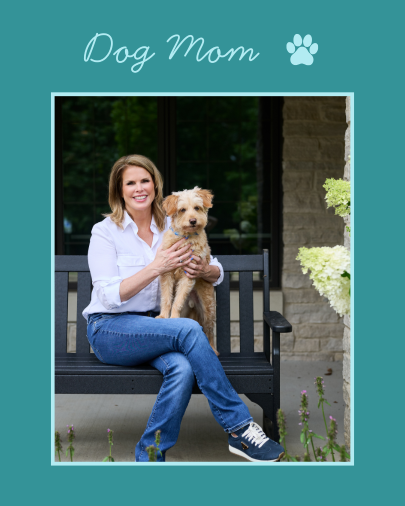 A woman sitting on a black bench outdoors, holding a small tan dog with fluffy fur. She is smiling and wearing a white shirt, blue jeans, and dark sneakers. The setting is beside a building with stone walls and some plants nearby.