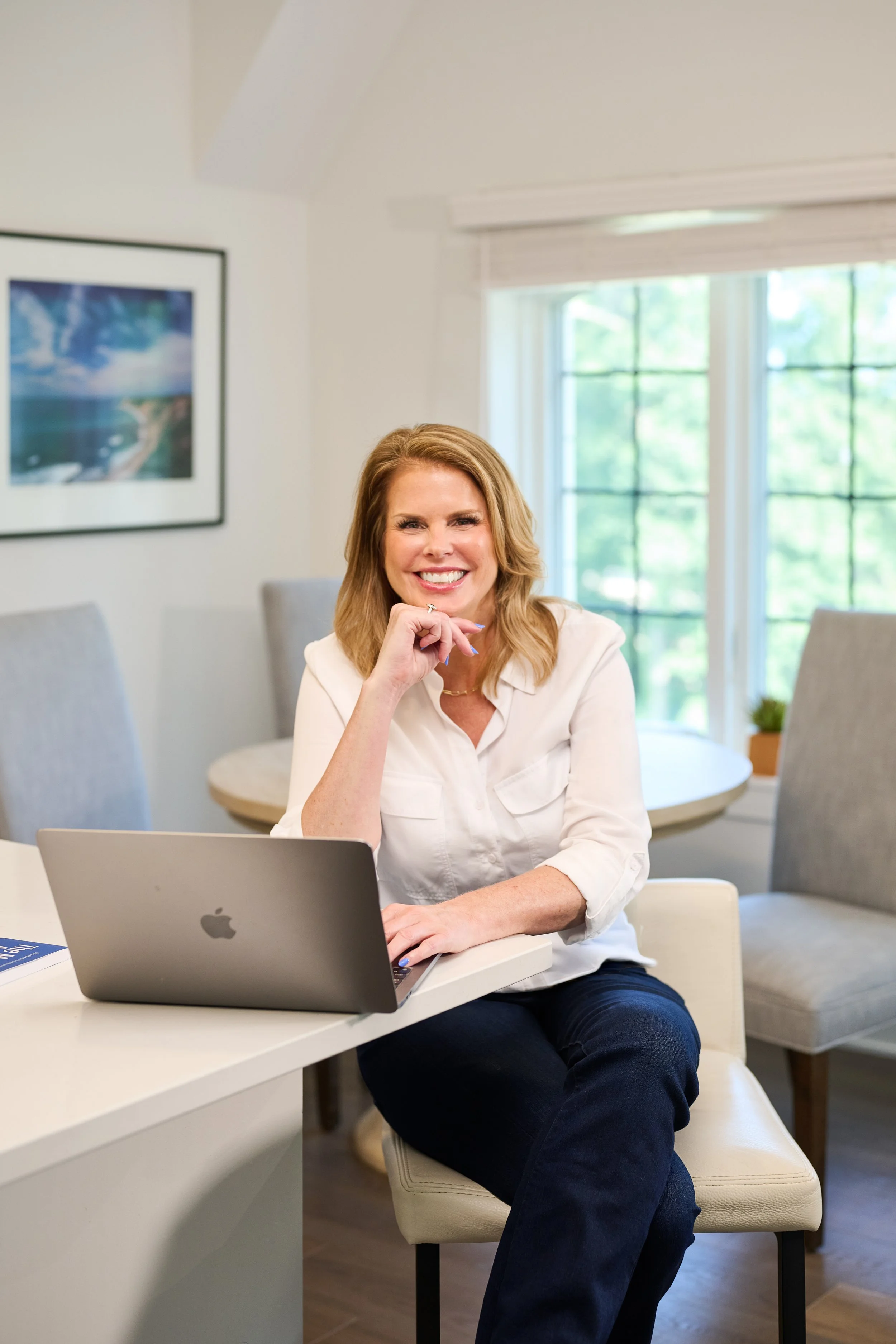 A woman with shoulder-length blonde hair, smiling and sitting at a white table in a bright room with large windows. She is wearing a white shirt and dark jeans, with a silver laptop in front of her.