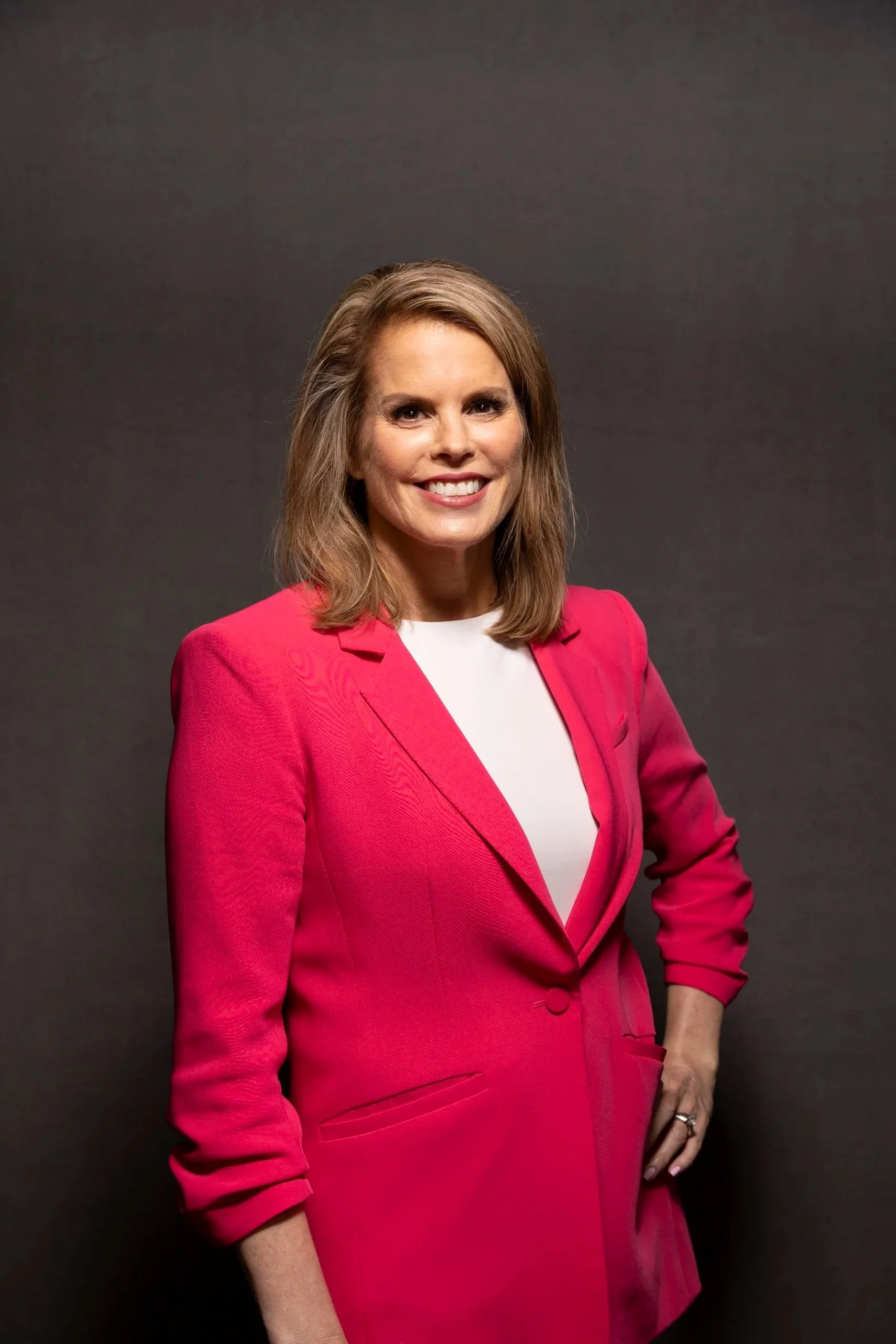 A woman with shoulder-length brown hair smiling, wearing a bright pink blazer over a white top, standing against a plain dark background.