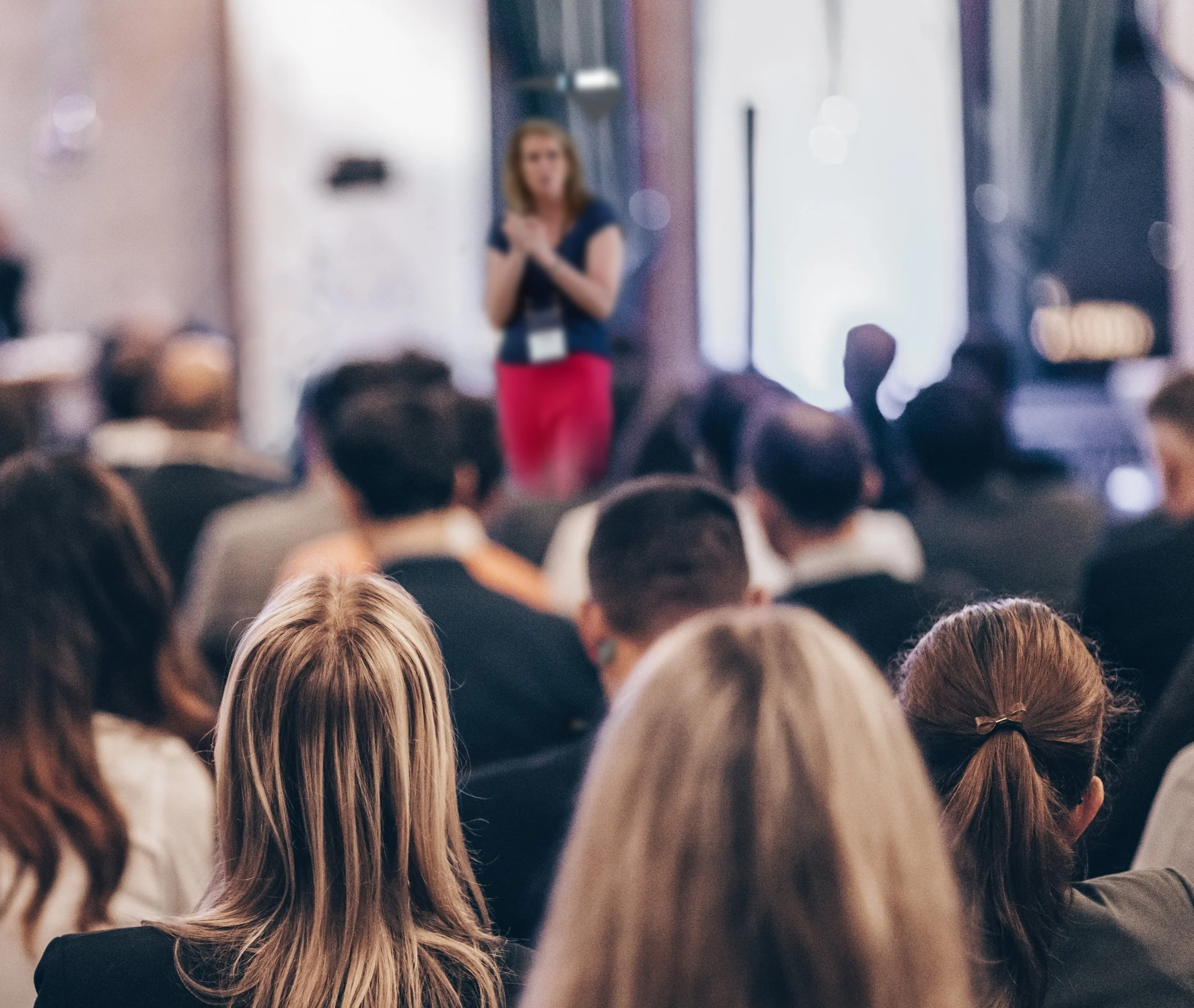 A woman with blonde hair, wearing a dark top and red pants, standing in front of a room full of seated audience members, speaking or presenting.