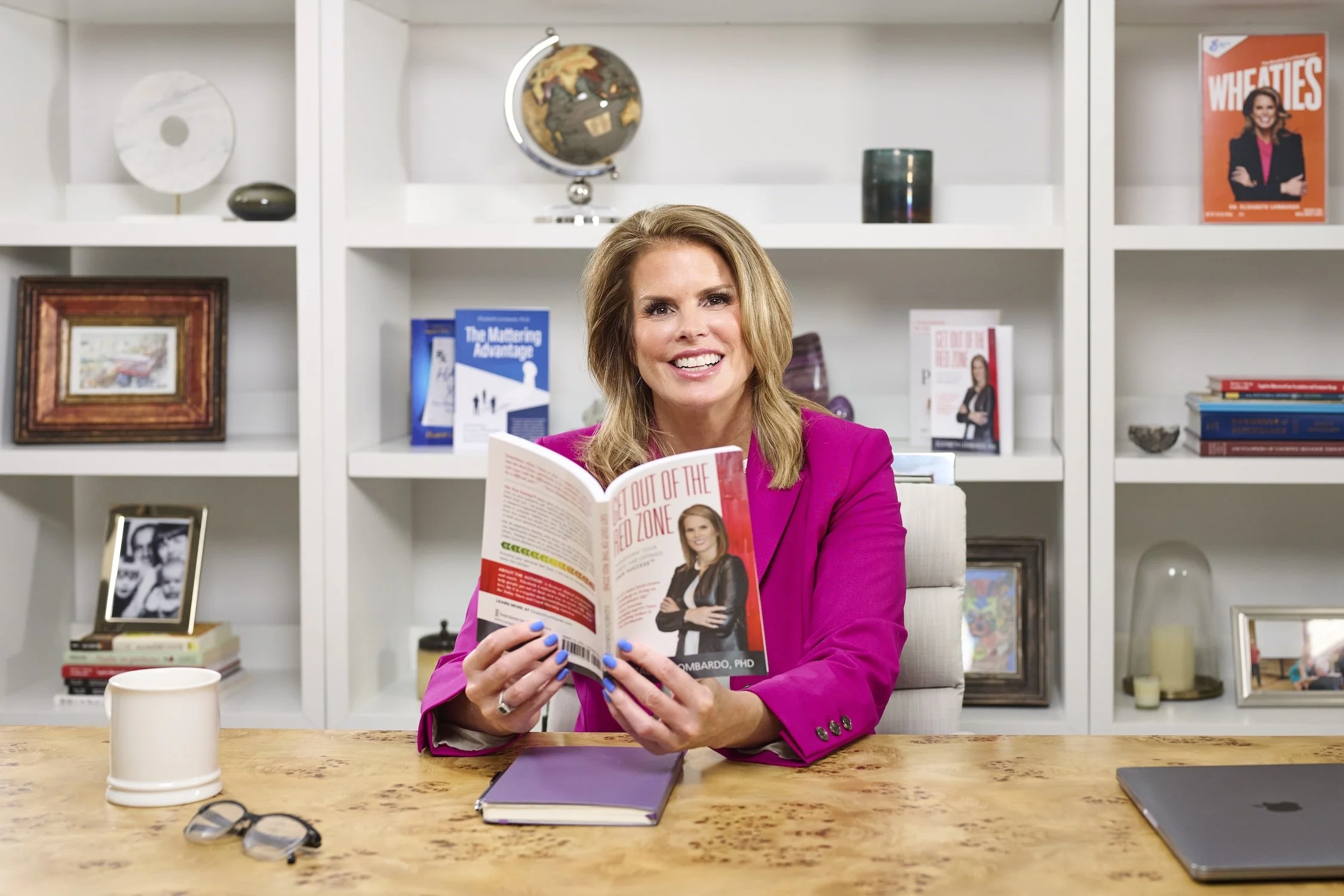 A smiling woman with blonde hair wearing a pink blazer is sitting at a wooden table, holding a magazine with a photo of herself on the cover. The background shows a white bookshelf with decorative items, books, and framed pictures.