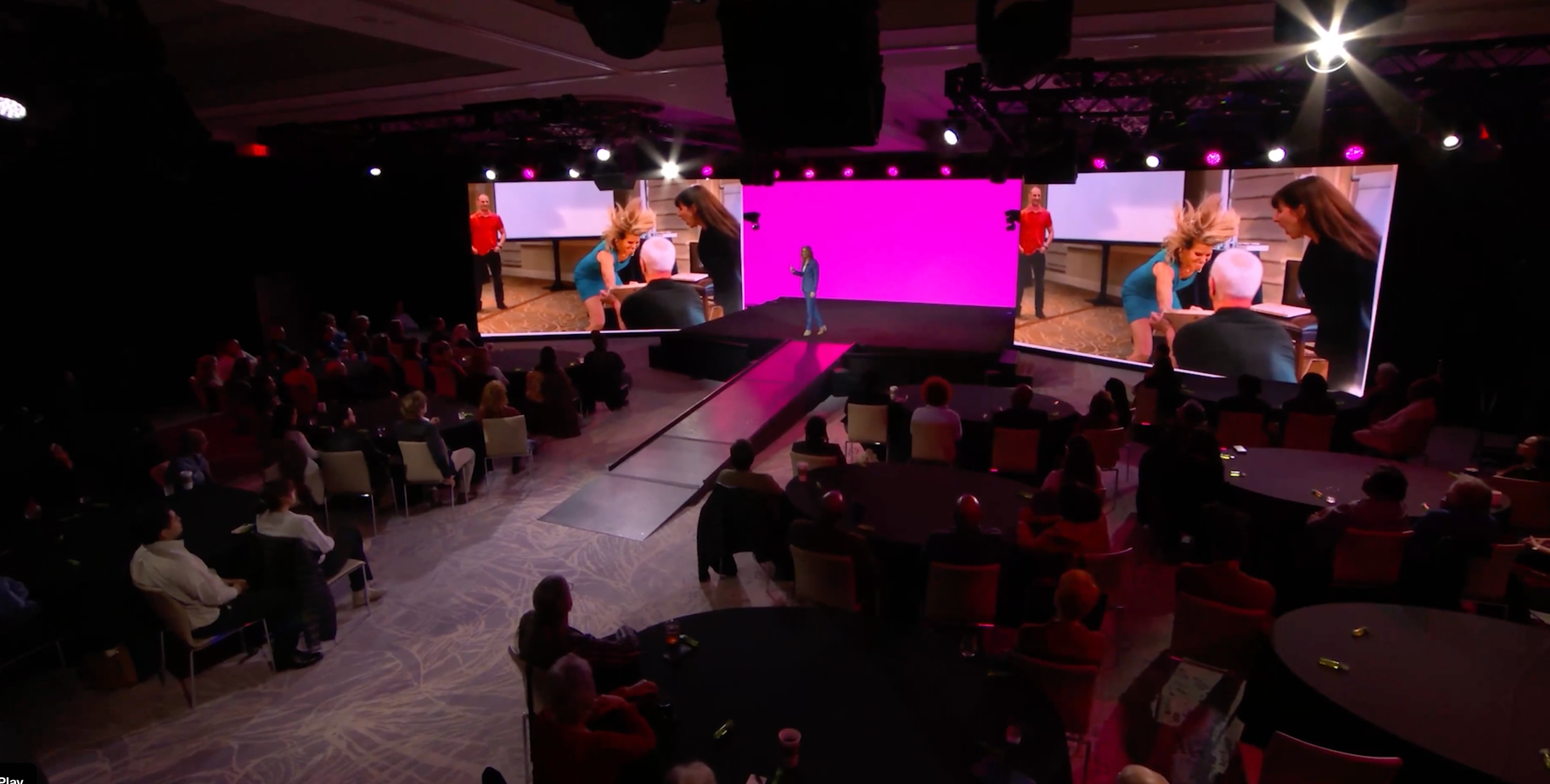 A large conference room with round tables filled with people watching a presentation on a stage. The stage features a woman speaking and a large screen displaying a pink background and images of people in a meeting.