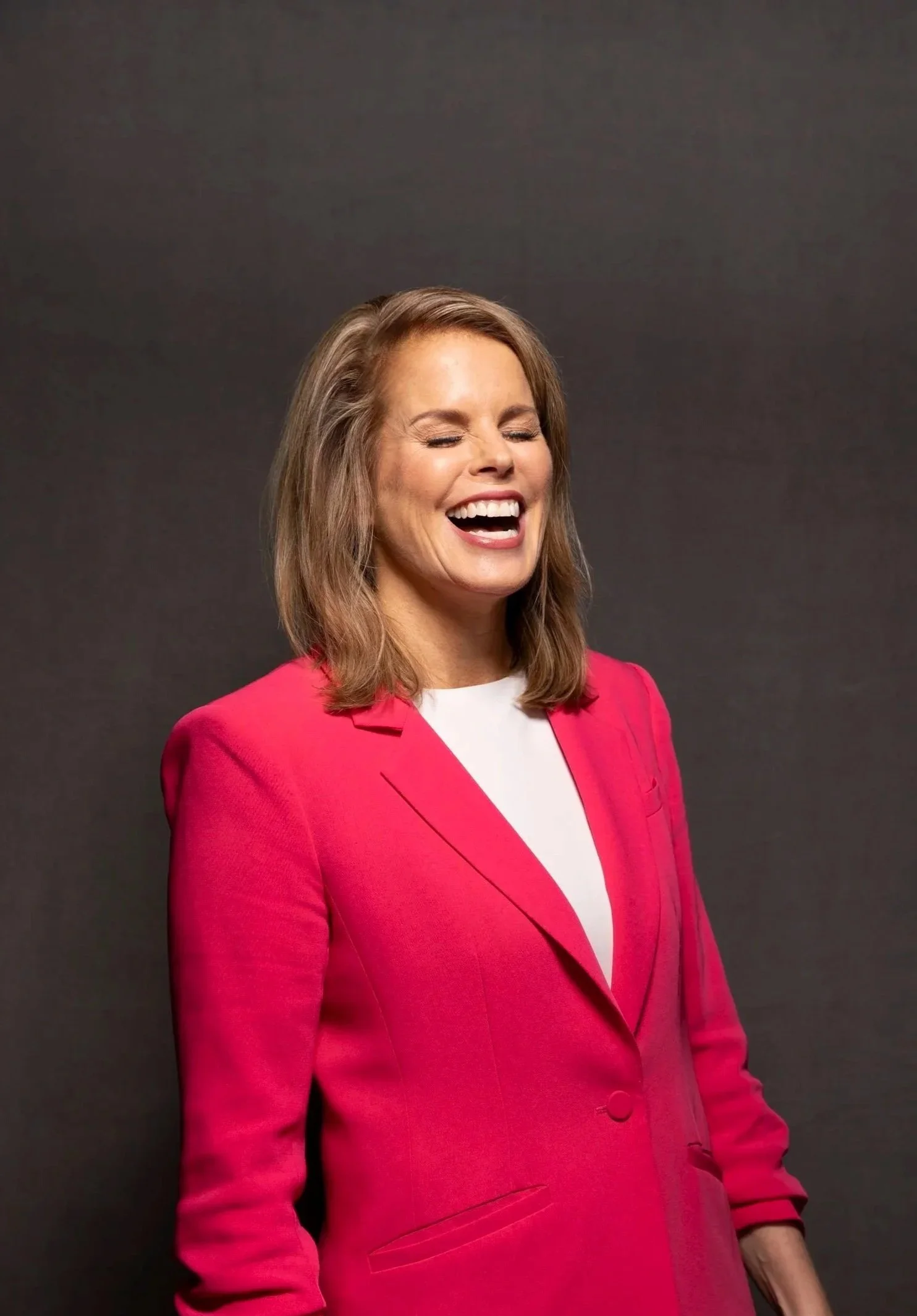 A woman with shoulder-length light brown hair, wearing a red blazer and white shirt, smiling with eyes closed against a plain dark background.