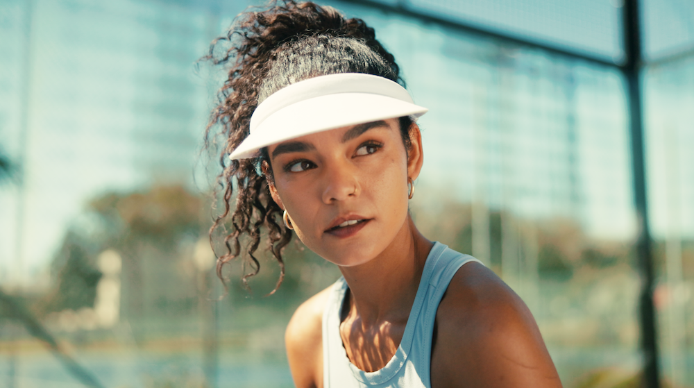 Young woman with curly hair in a ponytail wearing a white athletic visor, light blue tank top, and sports earrings, outdoors in a sunny area with glass buildings in the background.
