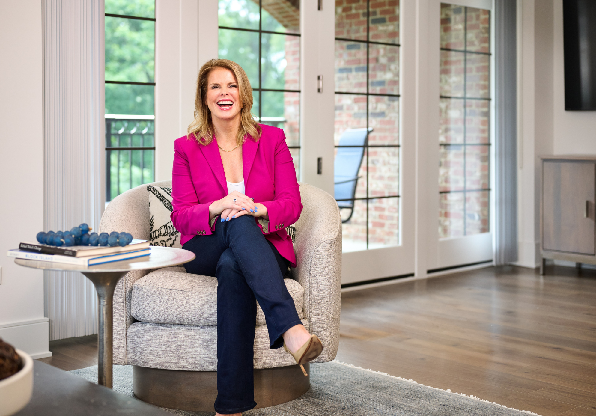 Woman with shoulder-length blonde hair wearing a bright pink blazer, white top, and dark jeans, sitting on a beige armchair, laughing in a modern living room with large windows, a brick wall, and a sliding glass door.