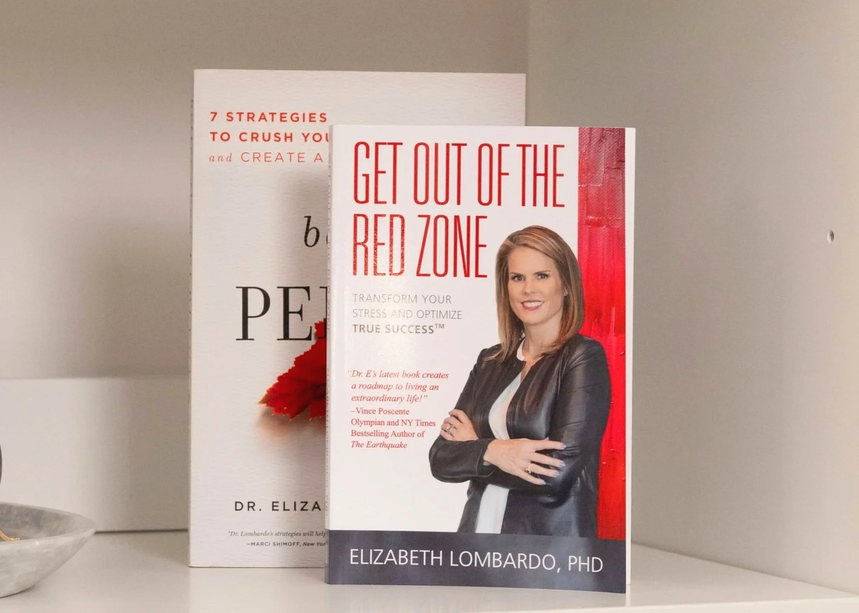 A book titled "Get Out of the Red Zone" by Elizabeth Lombardo, PhD, features a woman with shoulder-length brown hair, wearing a black leather jacket and white blouse, standing with arms crossed in front of a red curtain background. Behind the book, another similar book is partially visible.
