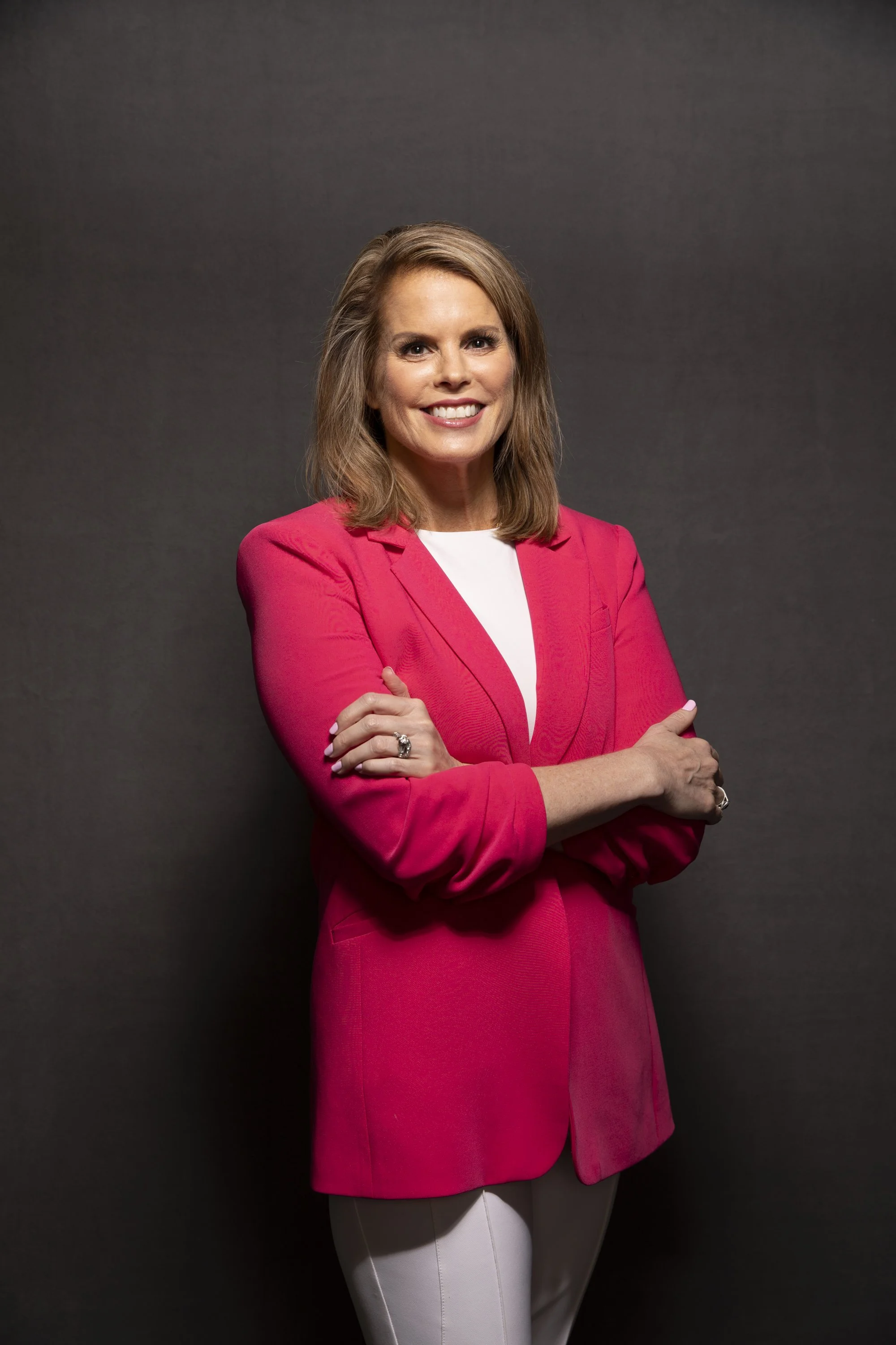 A woman with shoulder-length light brown hair smiling and crossing her arms, wearing a bright pink blazer over a white top, against a dark gray background.