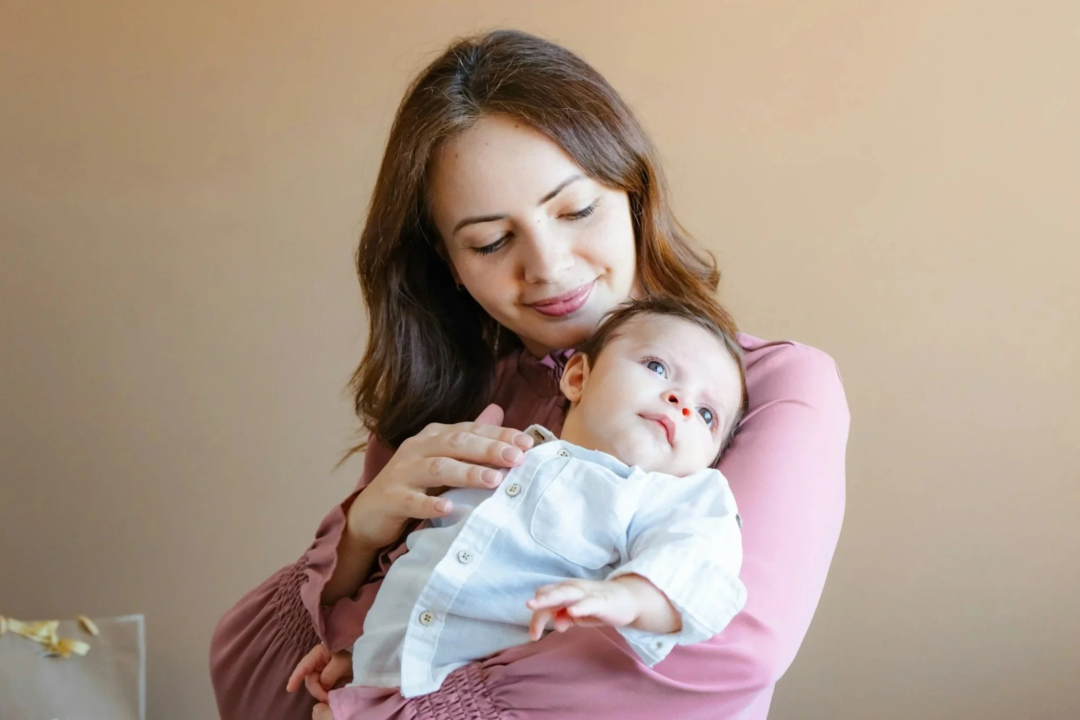 A woman with brown hair and fair skin holding a baby with blue eyes and light skin, both wearing light-colored clothing, standing against a beige wall.