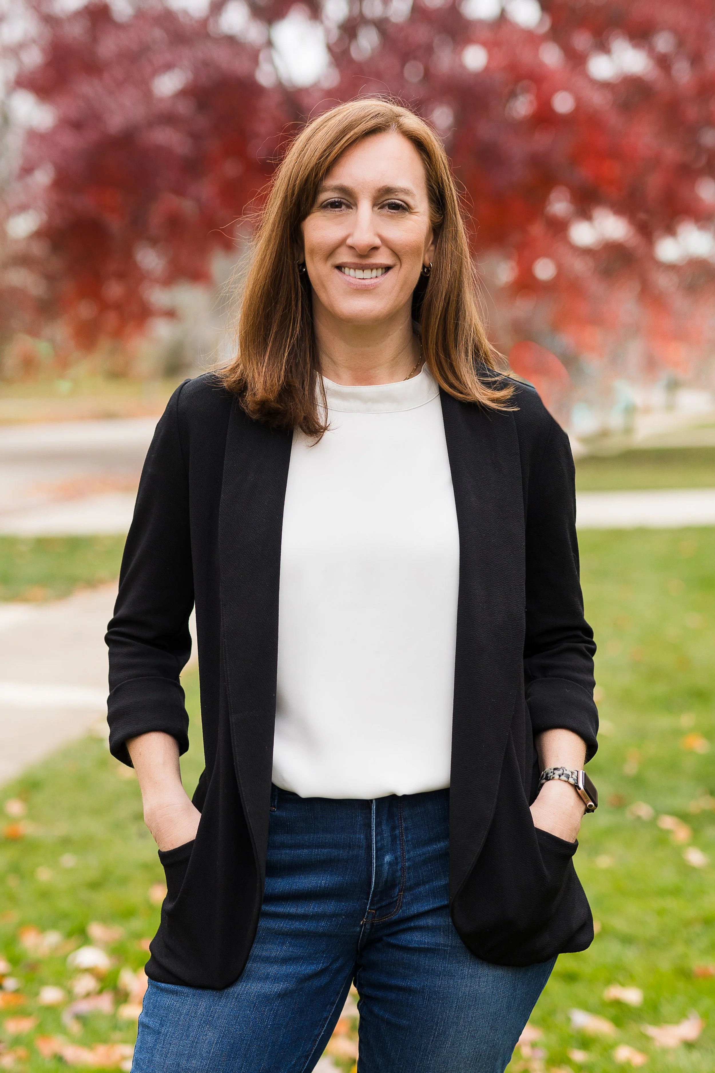 Dr. Tabatha Rienour is standing outdoors in front of a tree with red leaves, wearing a black blazer over a white top and jeans.