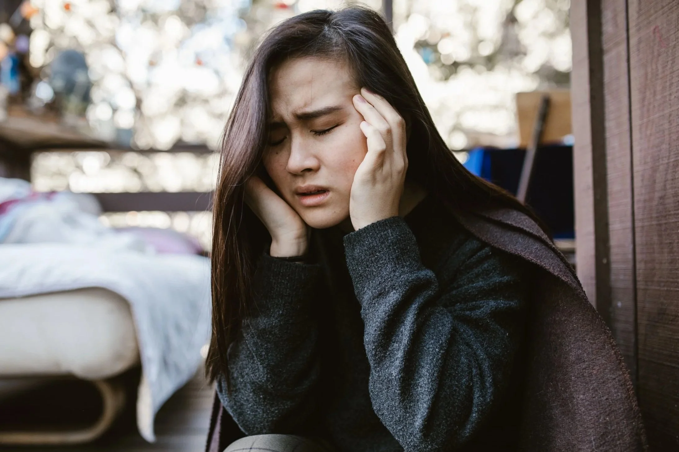 A young woman with long dark hair holding her head with closed eyes, appearing distressed or in pain, seated outdoors next to a brick wall. The image is meant to depict a person suffering from tinnitus.